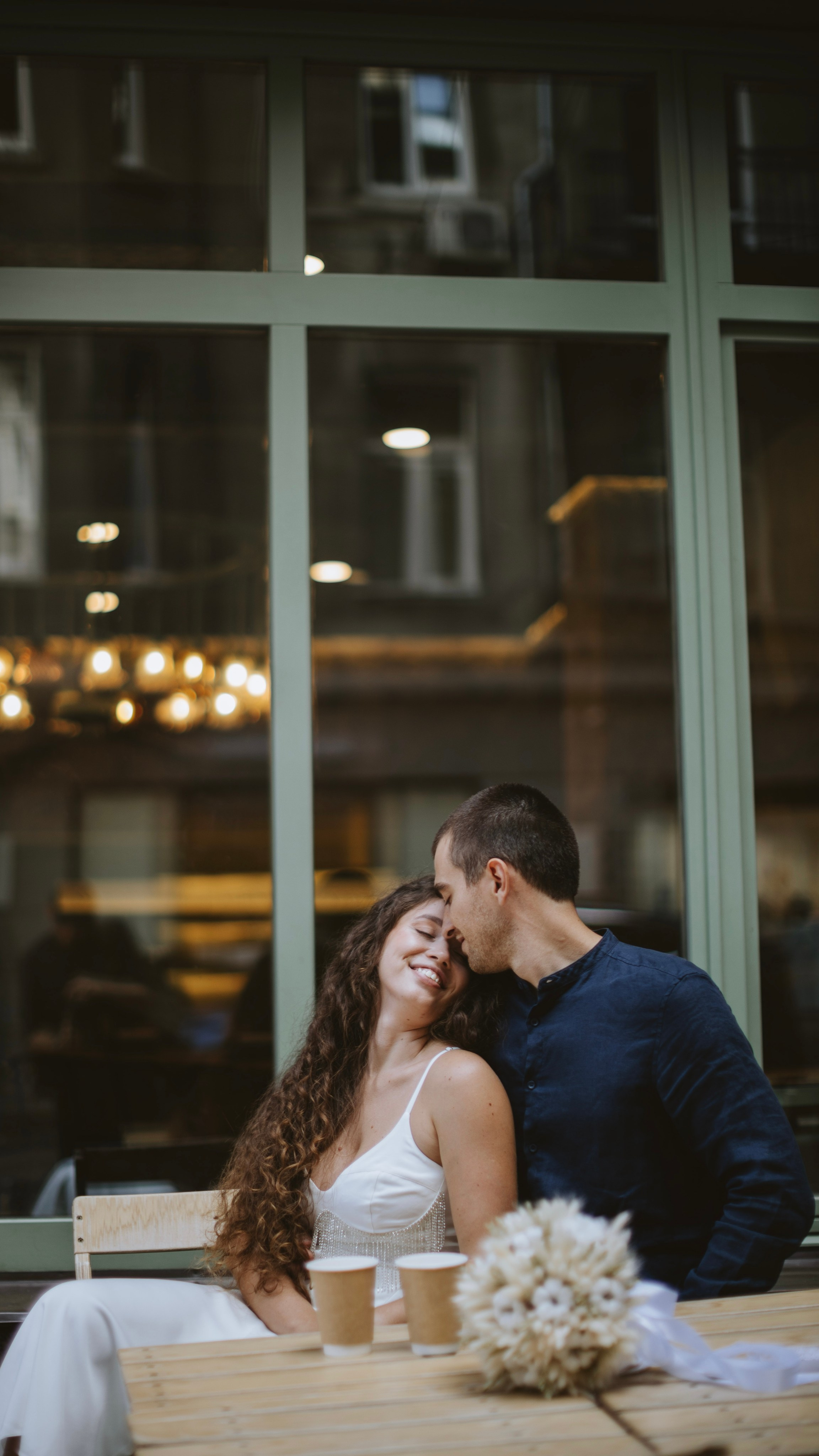 “Una coppia siede al tavolo di un caffè, condividendo un momento di intimità mentre sorride e si china l'uno verso l'altra, con i volti vicini. La donna, che indossa un semplice abito bianco con delicati ornamenti, tiene in mano un bouquet di fiori. Davanti a loro, due tazze di caffè sono appoggiate sul tavolo, con una luce morbida e calda dallo sfondo che si riflette attraverso la vetrata”.