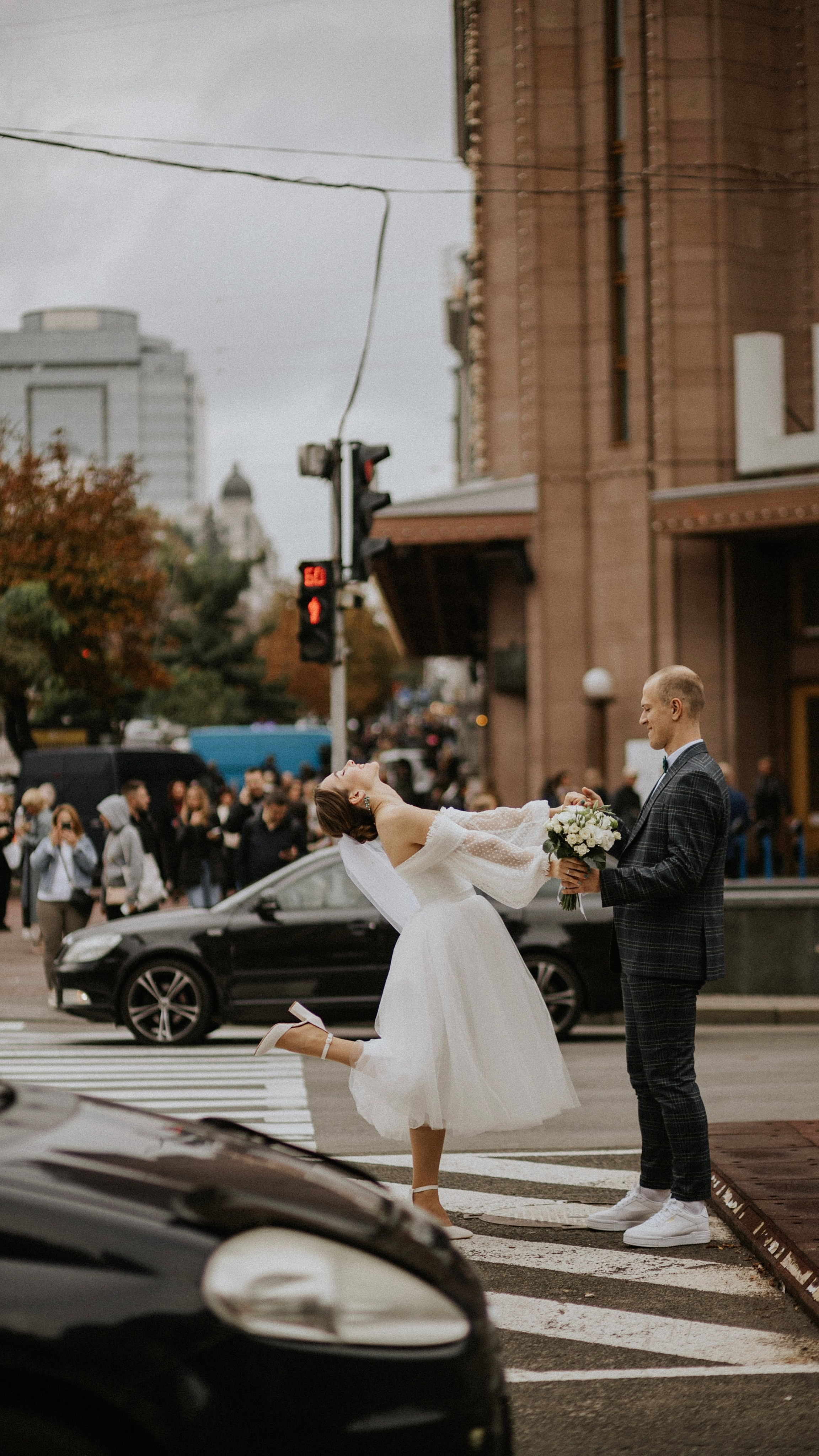 “Una sposa giocosa, vestita con un abito da sposa corto e fluente, gira gioiosamente sulle strisce pedonali con le braccia aperte e la testa inclinata all'indietro, con il velo che le ricade dietro. In una mano tiene un bouquet, mentre il suo compagno, vestito con un abito a scacchi e scarpe da ginnastica bianche, le sta di fronte con un sorriso gentile, tenendole l'altra mano. La strada trafficata e il traffico aggiungono uno sfondo dinamico al momento, pieno di divertimento e di festa”.