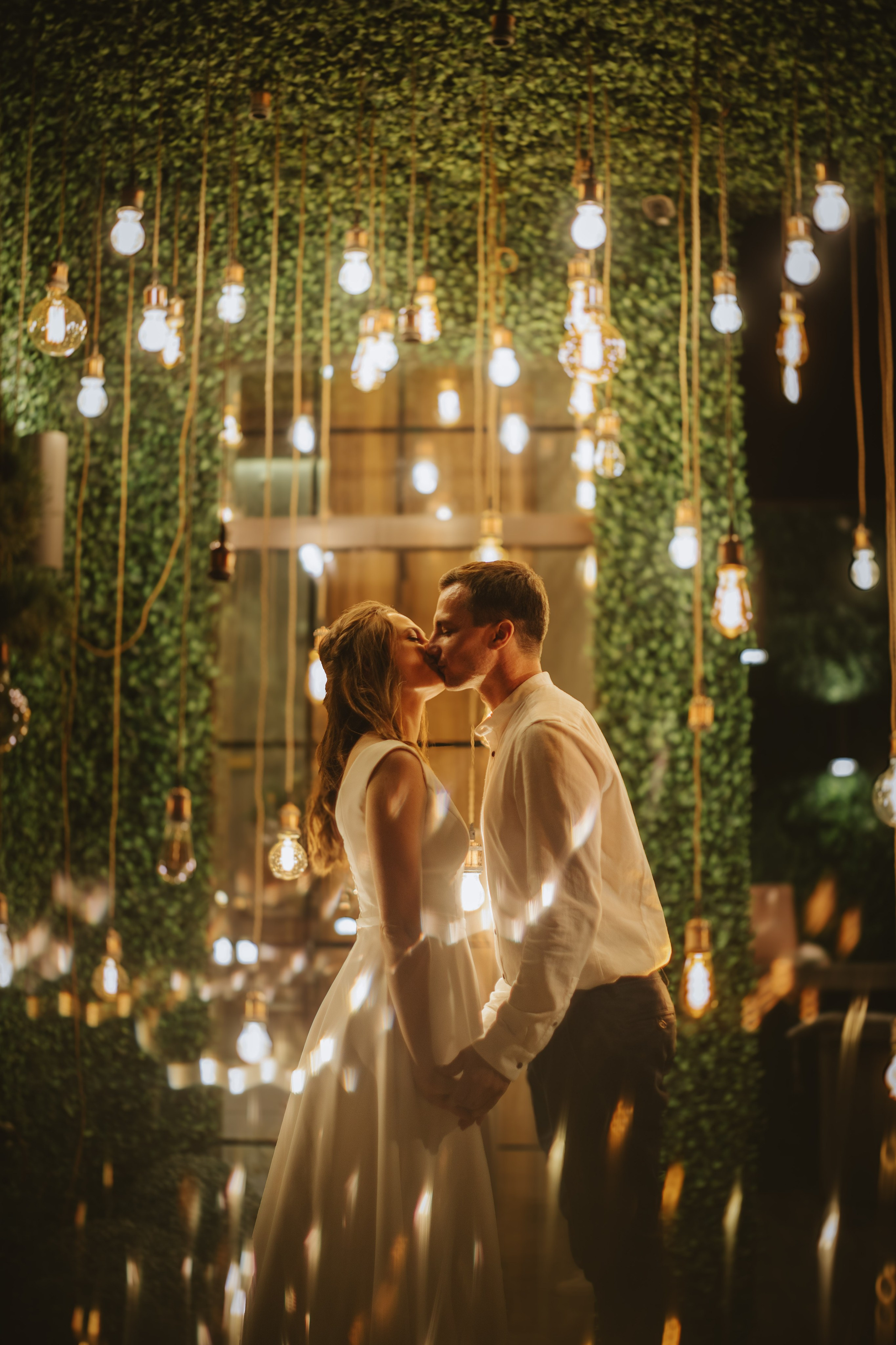 “Una coppia si scambia un bacio romantico davanti a uno sfondo splendidamente illuminato da lampadine sospese e verde lussureggiante. Le loro mani si stringono dolcemente mentre sono vicini, immersi nel caldo bagliore delle luci. I riflessi morbidi e l'illuminazione ambientale conferiscono al momento un'atmosfera intima e sognante”.