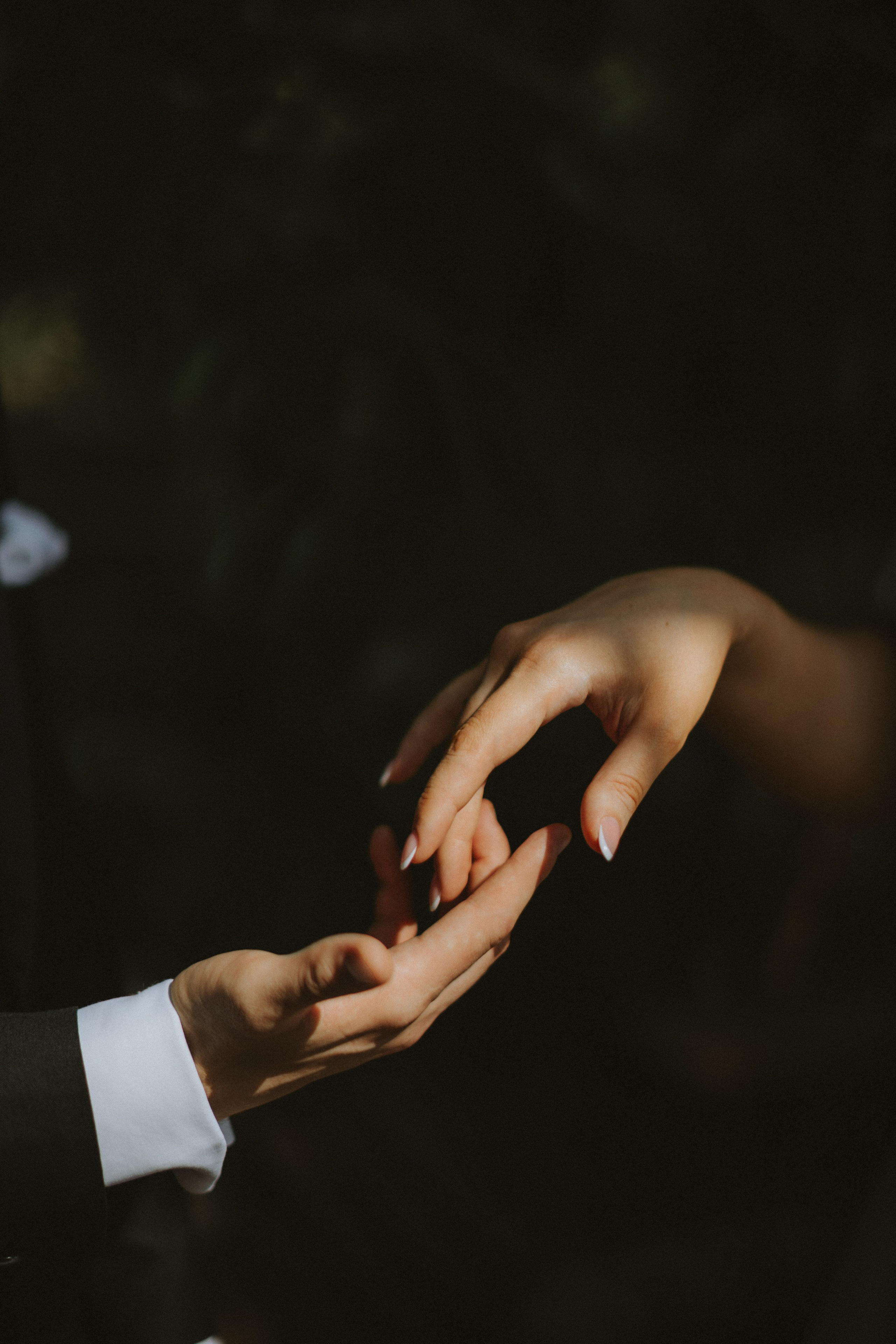 “Un'inquadratura ravvicinata delle mani di una coppia che si sfiorano delicatamente. Lo sposo, vestito con un abito nero e una camicia bianca, allunga la mano per incontrare quella della sposa, ornata da una delicata manicure francese. L'illuminazione proietta ombre morbide e mette in risalto il loro intimo legame su uno sfondo scuro e sfocato, creando un momento romantico e intimo”.