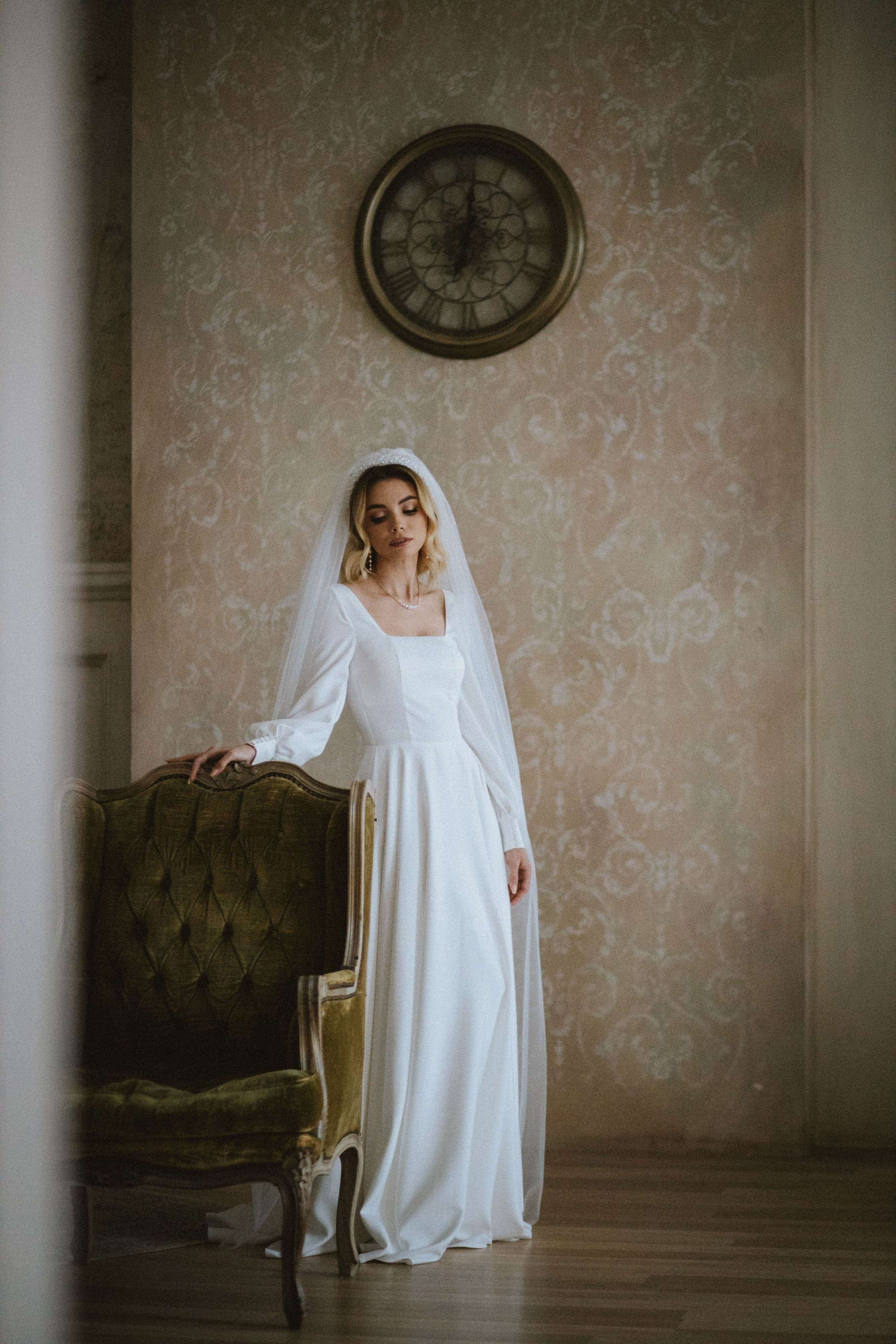 “Una sposa in un semplice ma elegante abito bianco con maniche lunghe e velo, in piedi accanto a un'antica poltrona verde. Guarda pensierosa, con una postura calma e aggraziata, mentre alle sue spalle è appeso alla parete un orologio d'epoca”.