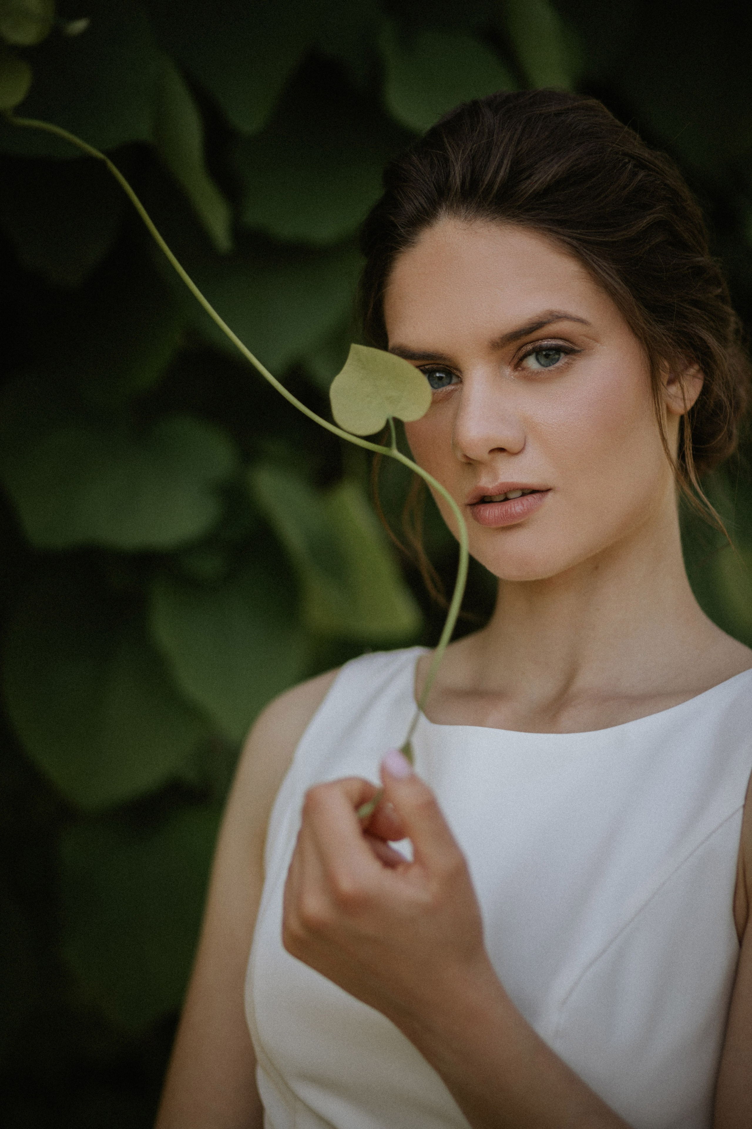 “Una sposa in un abito bianco minimalista si staglia con grazia su uno sfondo verde e rigoglioso. Tiene una vite con una singola foglia vicino al viso, guardando direttamente l'obiettivo con un'espressione serena”.