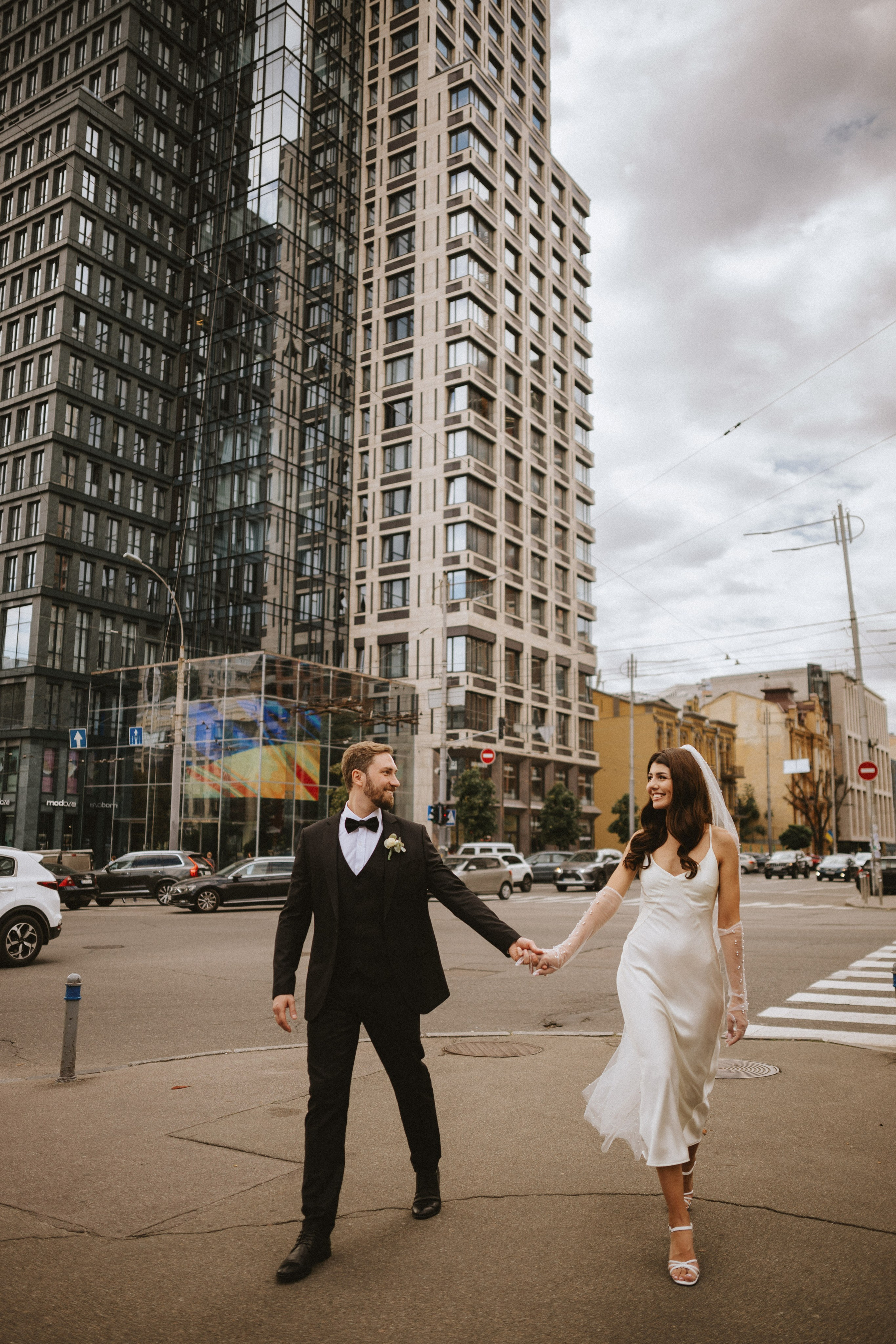 “La sposa e lo sposo camminano mano nella mano per le strade della città, sorridendosi l'un l'altro sullo sfondo dell'architettura moderna. La sposa, con il suo elegante abito da sposa e il velo, irradia eleganza, mentre lo sposo appare elegante nel suo smoking nero, catturando un perfetto momento di romanticismo urbano”.