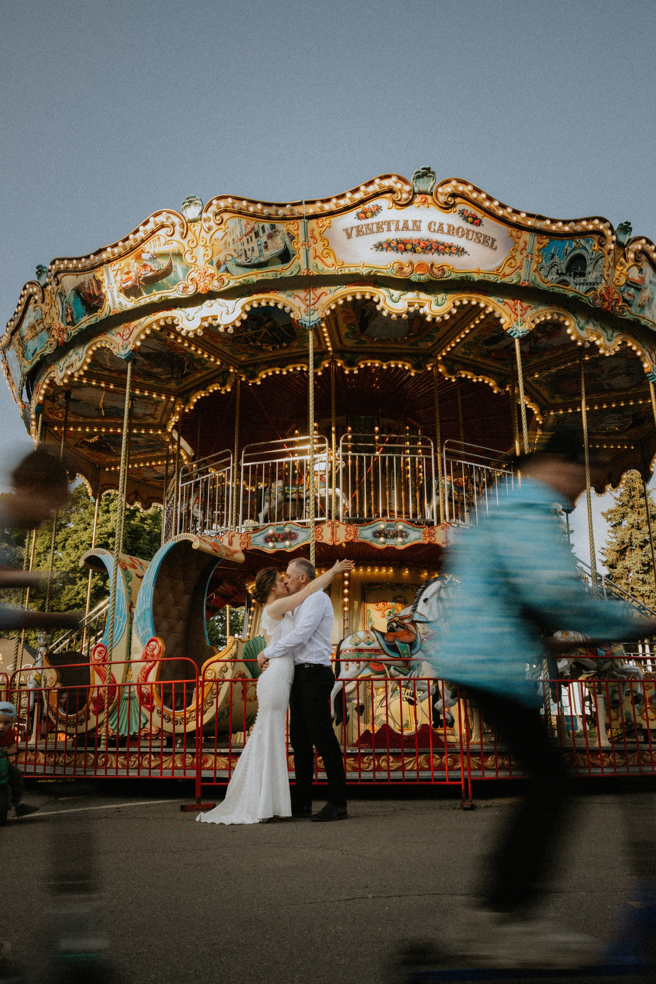 “Una coppia si scambia un bacio davanti a una vivace giostra veneziana; il loro momento di gioia è catturato sullo sfondo di luci colorate. La sposa, in un elegante abito bianco, abbraccia lo sposo, mentre la sfocatura del movimento di un passante aggiunge una dinamica interessante alla scena”.