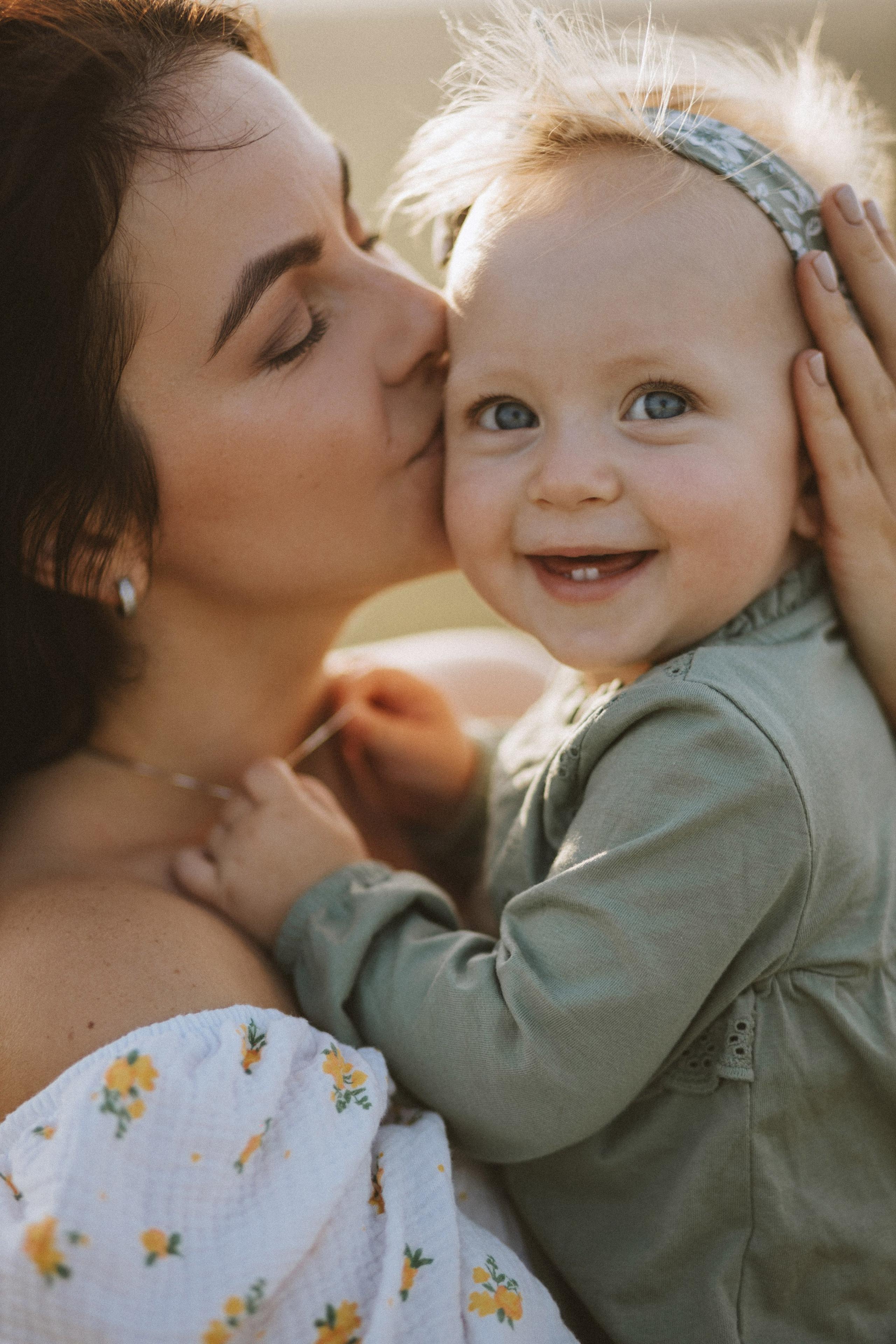 “Un gioioso primo piano di una madre che bacia la sua sorridente figlioletta, che indossa un vestitino verde e una simpatica fascia per capelli. Gli occhi blu della bambina brillano di felicità mentre tiene la mano della madre, con uno sfondo morbido e naturale”.