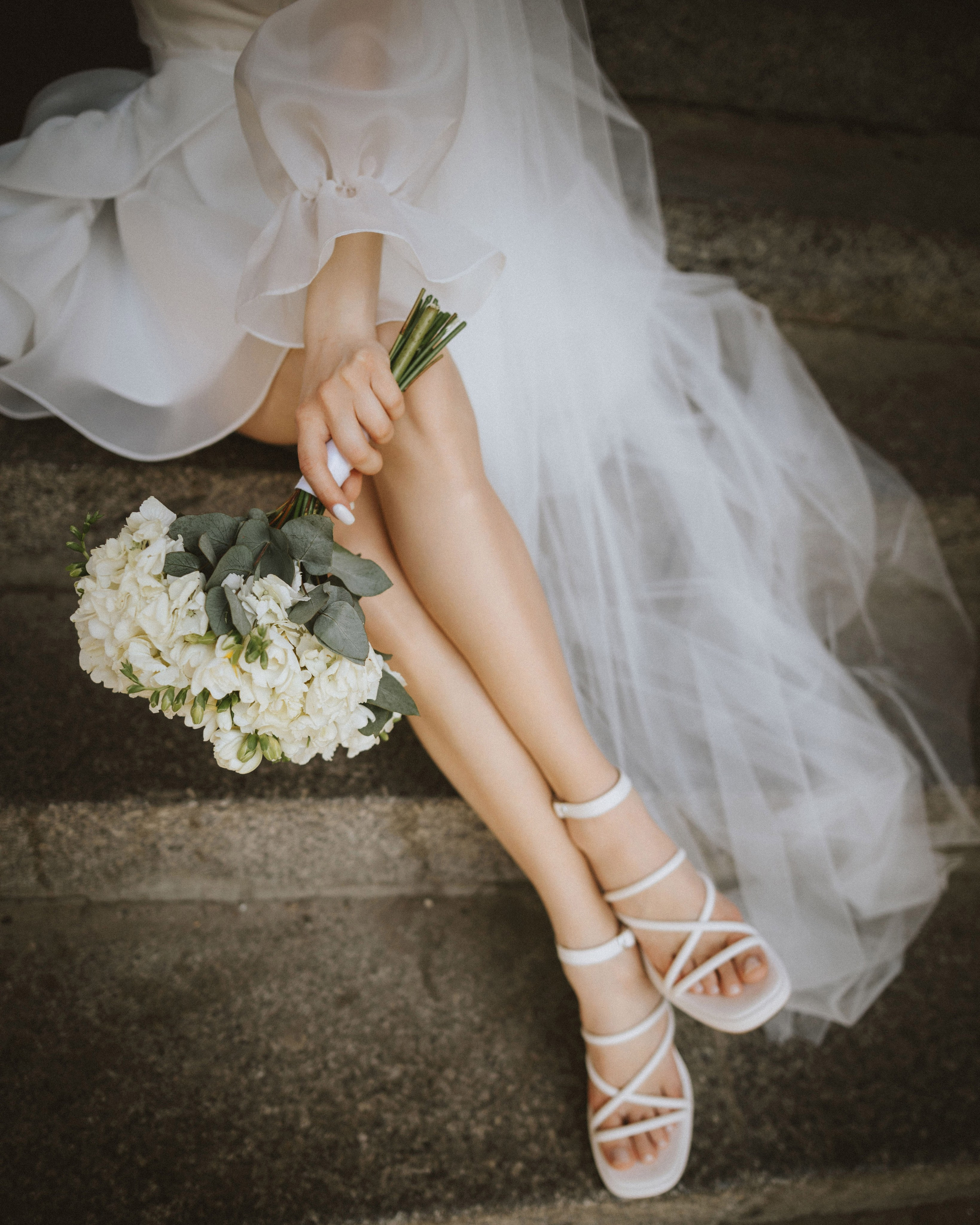 “Primo piano delle gambe di una sposa seduta su un gradino di pietra, che indossa un abito da sposa bianco e sandali bianchi. In una mano tiene un bouquet di fiori bianchi con foglie verdi, mentre l'altra mano è appoggiata sul grembo”.
