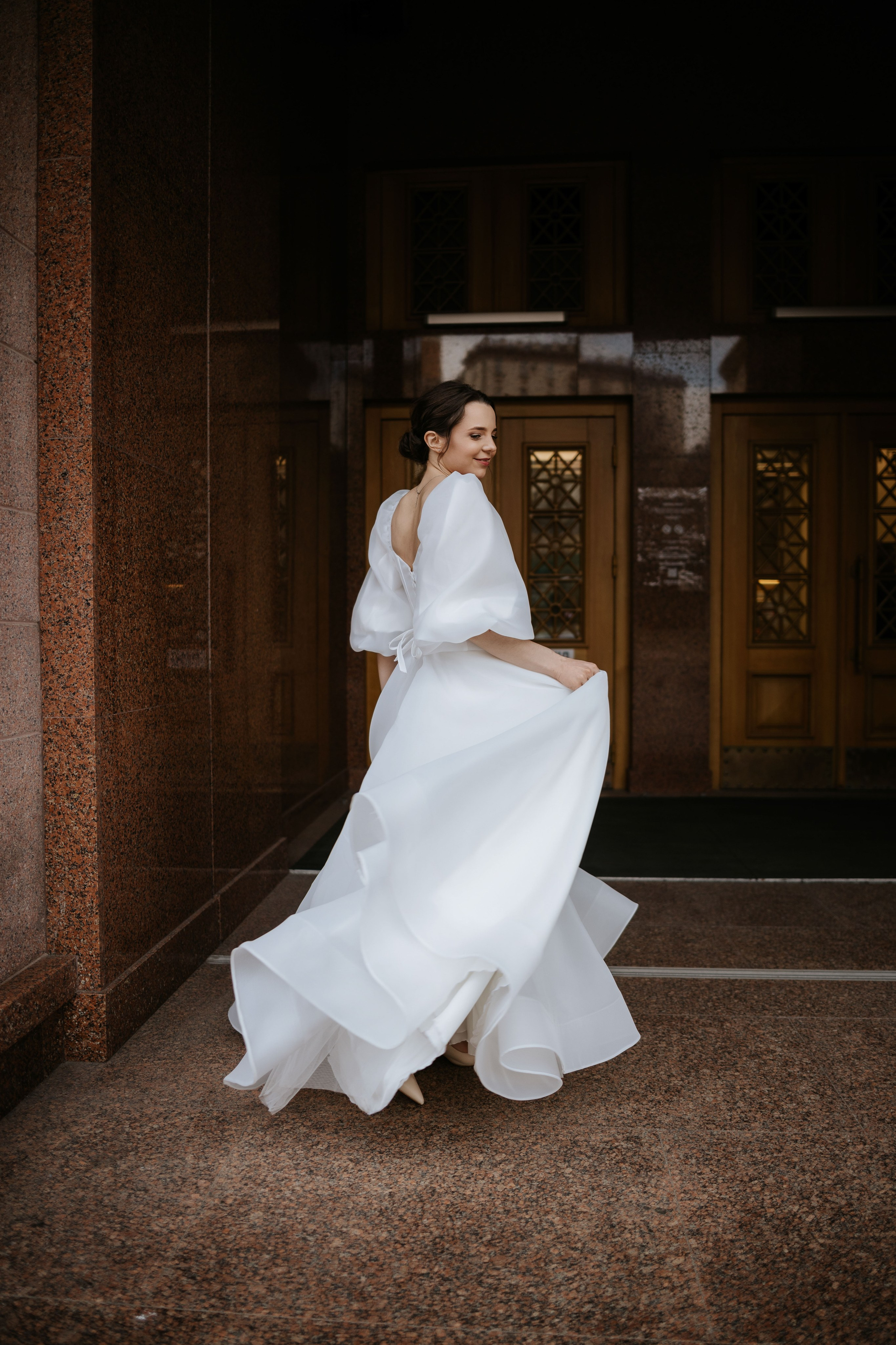“Una sposa dal sorriso radioso, che volteggia nel suo fluente abito bianco con maniche a sbuffo e schiena bassa, in piedi davanti a un grande edificio con eleganti porte di legno. Il movimento del suo abito crea pieghe morbide e aggraziate mentre ruota. La foto cattura il suo momento di gioia, evidenziato dalla luce naturale”.