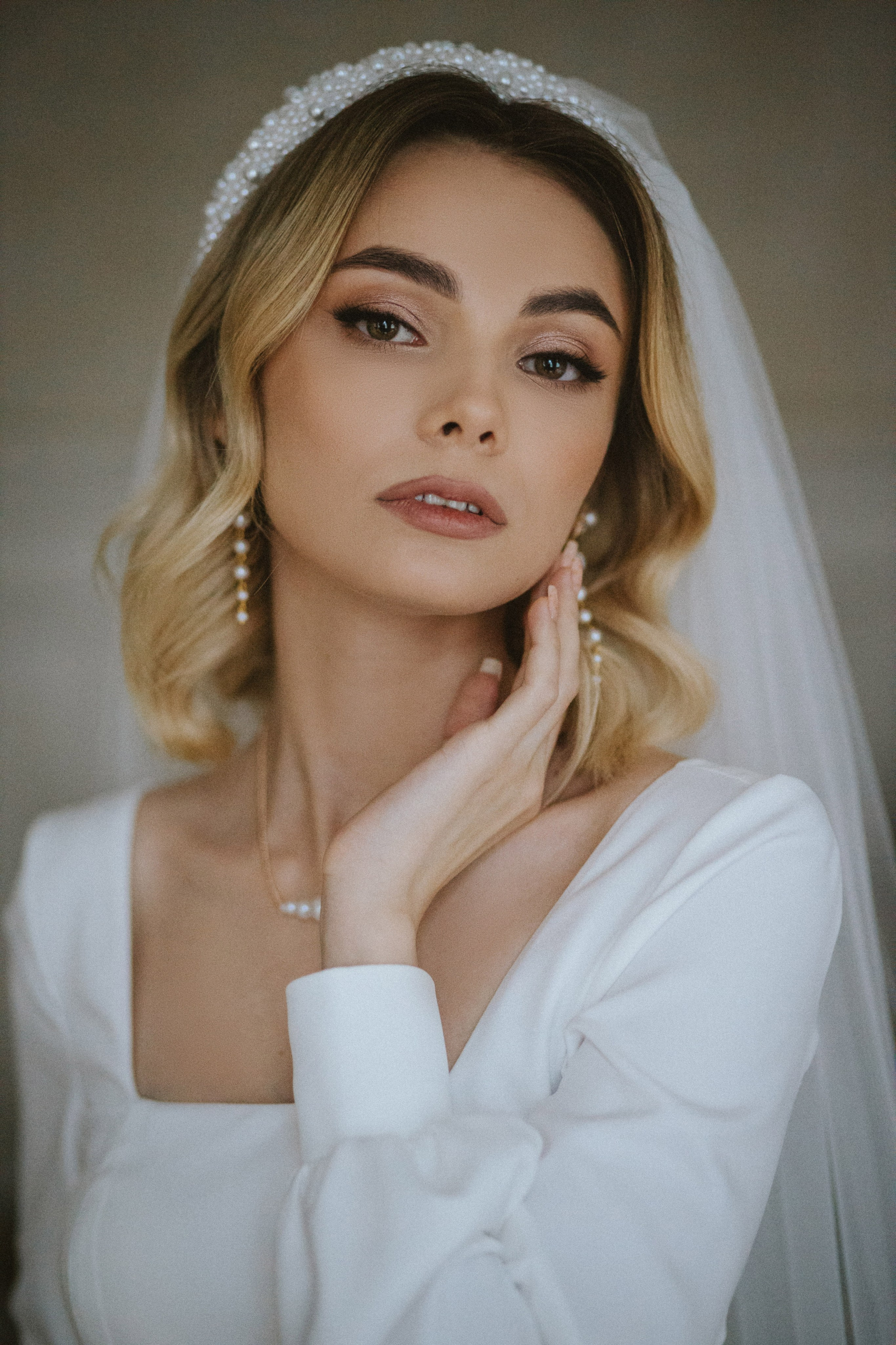 “Una sposa in posa con un'espressione morbida ed elegante. Si tocca delicatamente il viso con una mano mentre l'altra è appoggiata al fianco. La sposa indossa un cerchietto impreziosito da perle e orecchini delicati, mentre il suo abito bianco presenta maniche lunghe con scollatura quadrata”.