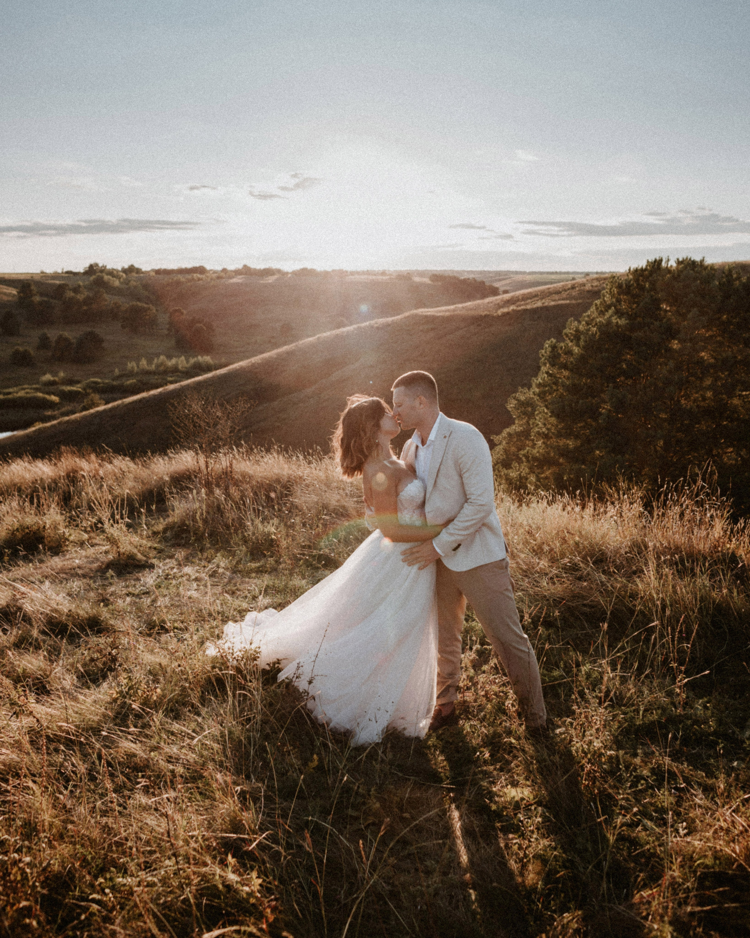 “Una coppia condivide un tenero bacio mentre si abbraccia sulla cima di una collina durante l'ora d'oro. La calda luce del sole inonda la scena, creando un'atmosfera romantica e sognante. L'abito della sposa scorre dolcemente intorno a lei, catturando la luce mentre sta vicino al suo partner. Le dolci colline e il verde lussureggiante sullo sfondo completano il paesaggio pittoresco, aggiungendo intimità al momento”.