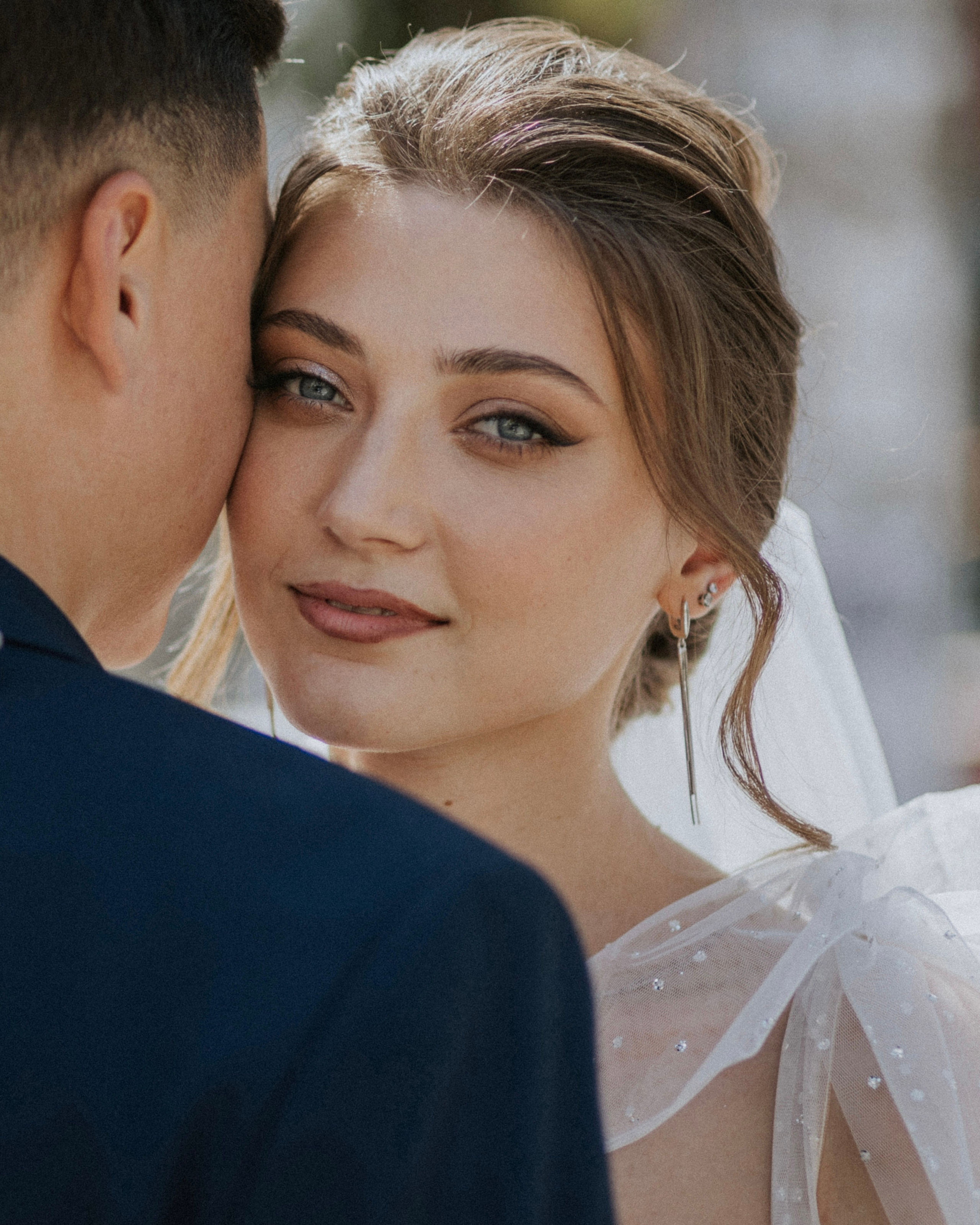 “Un primo piano di una sposa con un trucco morbido e naturale e orecchini delicati, che guarda amorevolmente la macchina fotografica. Indossa un abito da sposa con tessuto trasparente e scintillante sulle spalle. Lo sfondo è delicatamente sfocato, per concentrarsi sulla sua espressione serena e sul dolce legame con il suo partner, leggermente visibile sul lato sinistro dell'inquadratura”.