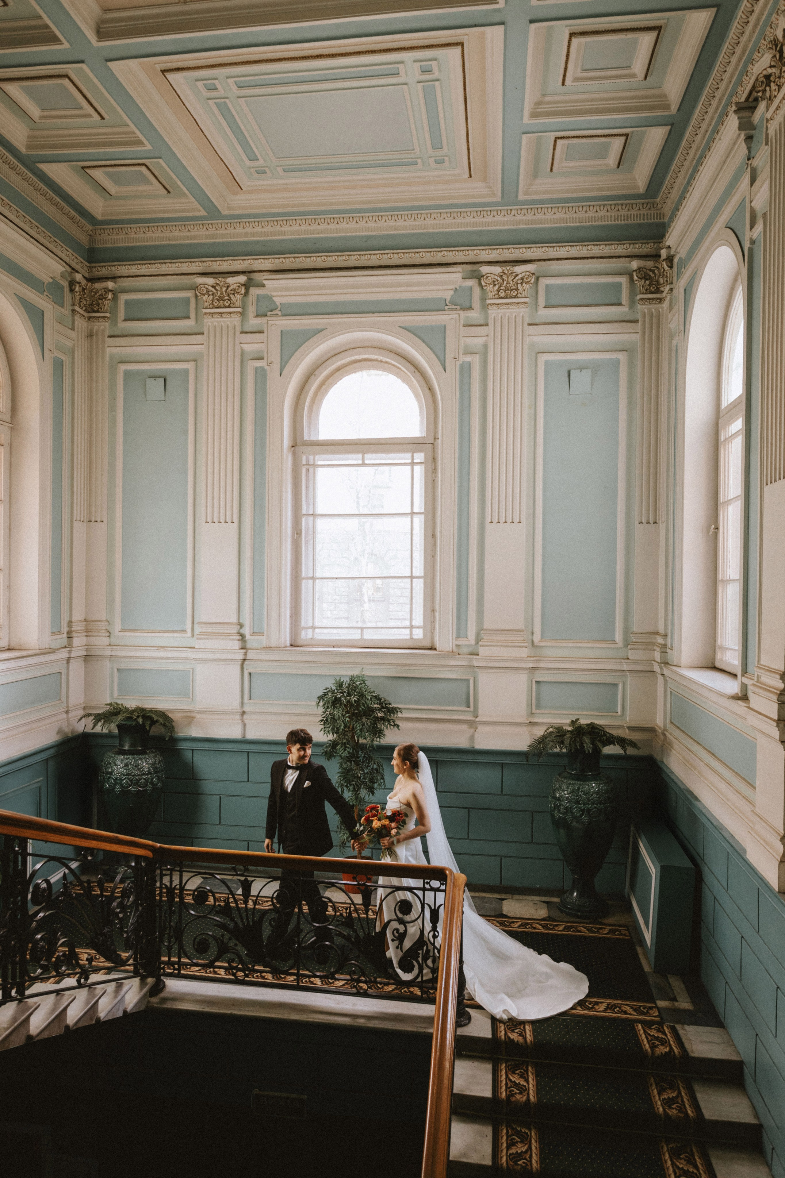 “Una coppia che cammina mano nella mano lungo una scalinata di un grande edificio classico. La sposa indossa un abito da sposa fluente e porta un bouquet, mentre lo sposo è vestito con un classico smoking nero. Le eleganti pareti blu e le grandi finestre riempiono lo spazio di luce naturale, aggiungendo un'atmosfera romantica”.