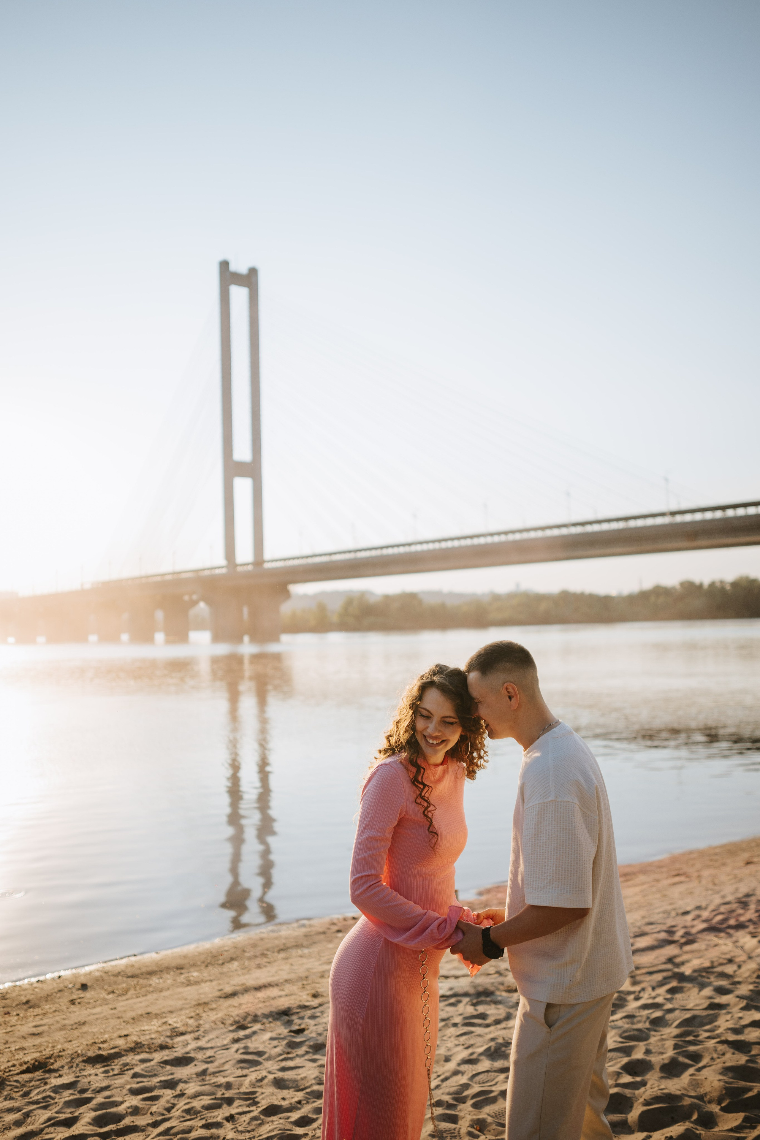 “Una coppia che si tiene per mano e si sorride, in piedi sulla sabbia vicino a un fiume al tramonto. La donna indossa un abito rosa e l'uomo una camicia bianca. Sullo sfondo, un ponte attraversa il fiume, illuminato dalla tenue luce dorata del sole al tramonto”.