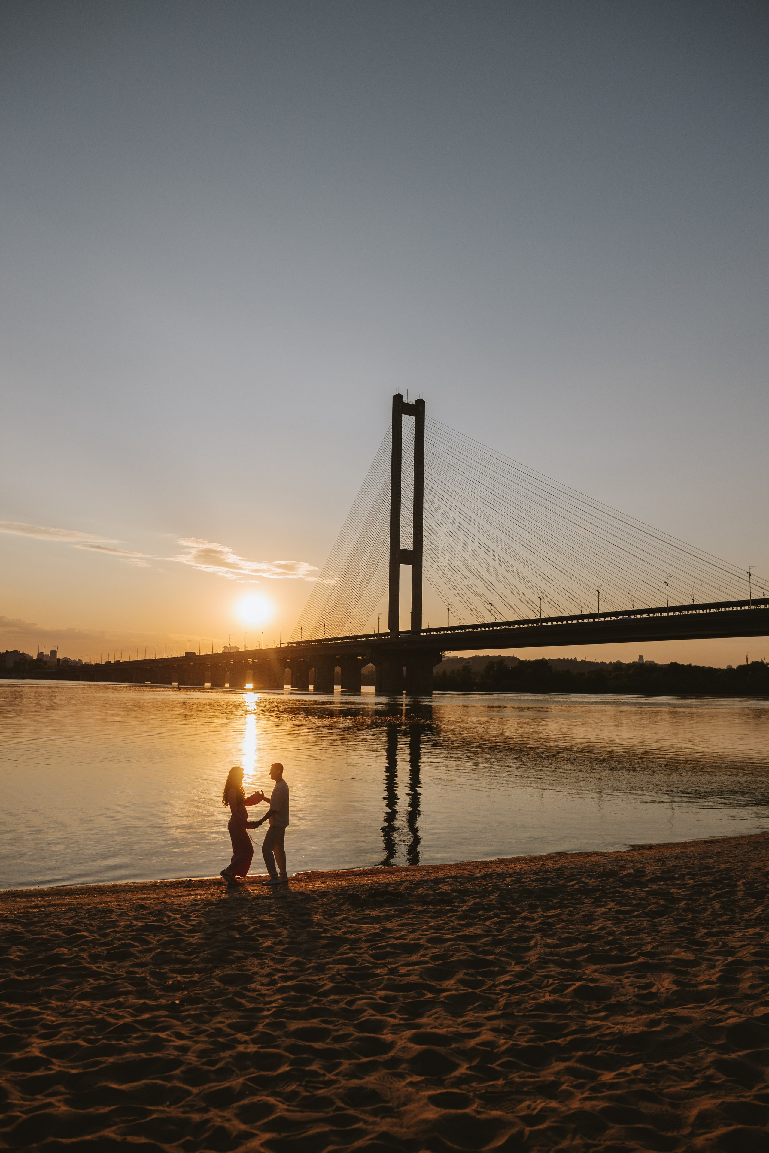 “Una coppia che si tiene per mano e balla sulla riva sabbiosa durante il tramonto, con un ponte che si staglia contro il cielo e il suo riflesso che brilla sull'acqua”.