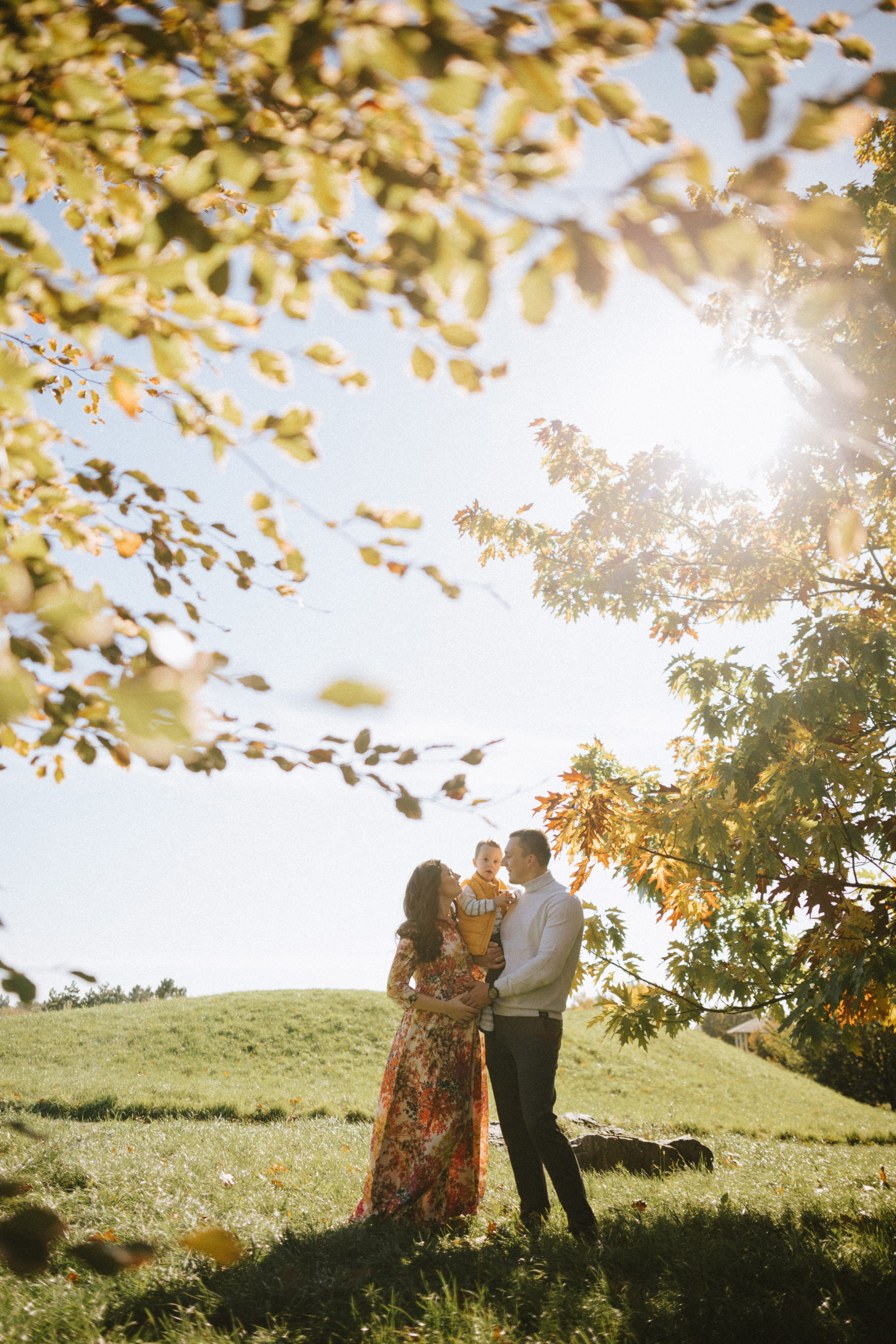 “Una famiglia felice di tre persone, in piedi all'aperto in un parco con fogliame autunnale. La madre tiene in braccio il figlio piccolo mentre il padre li abbraccia entrambi. La luce del sole filtra attraverso gli alberi, creando un'atmosfera calda e gioiosa”.