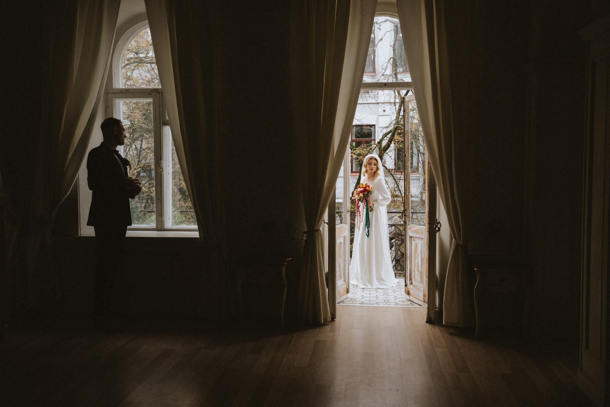“A bride in a white wedding dress holding a bouquet of vibrant flowers, standing at the entrance of a room, framed by open doors with soft curtains. Her partner is silhouetted against the light, standing near a window and looking at her, capturing a beautiful, intimate moment.”