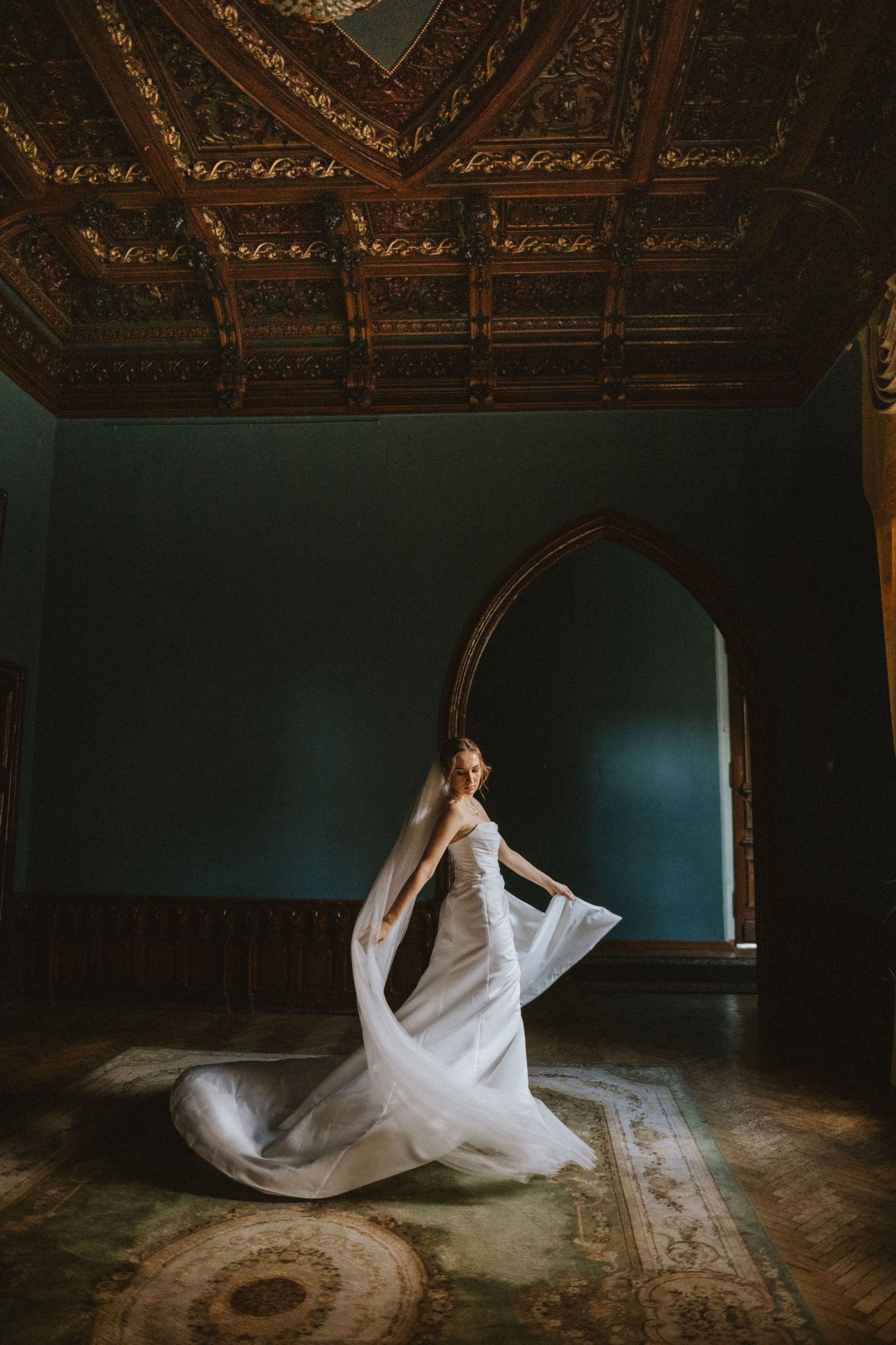 “Sposa elegante in un fluente abito bianco e velo in piedi in una grande sala d'epoca con soffitto in legno ornato e pareti blu scuro, tenendo il tessuto del suo abito mentre si drappeggia sul pavimento”.