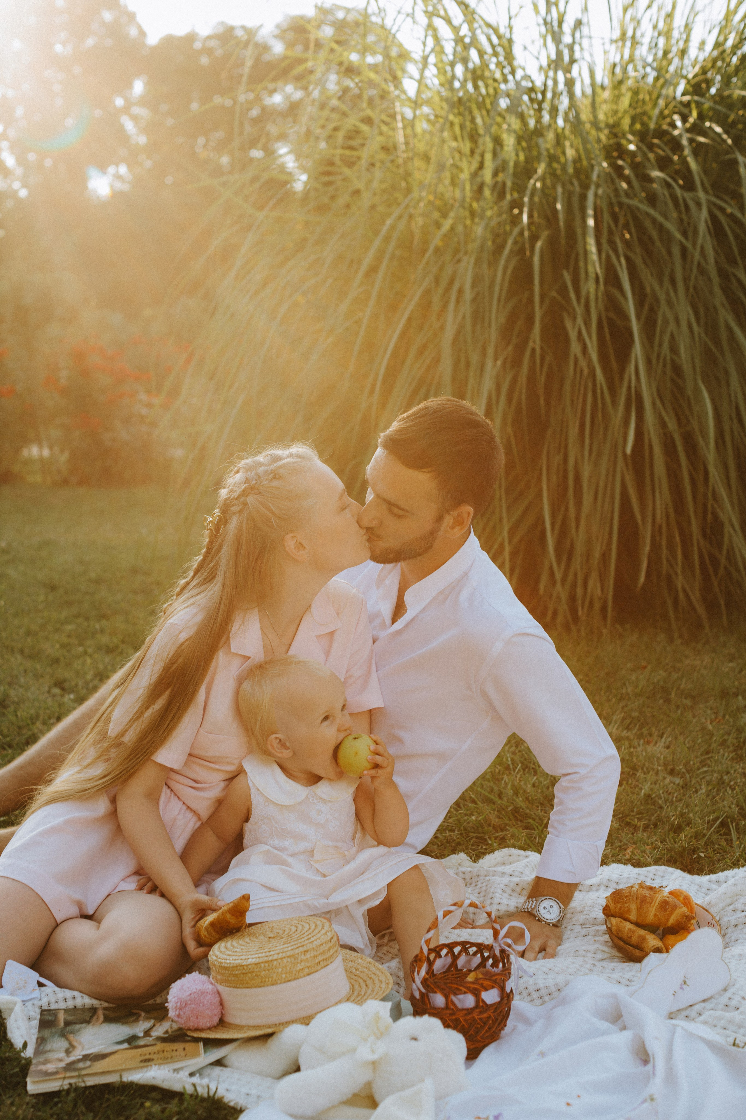 “Una famiglia si gode un tranquillo picnic all'aperto, immortalato dalla calda luce del sole. I genitori si scambiano un dolce bacio mentre il loro piccolo, seduto tra loro, sgranocchia una mela. La madre indossa un morbido abito rosa, mentre il padre è vestito con una camicia bianca. L'allestimento del picnic comprende un cappello di paglia, croissant e un cestino, con un'accogliente coperta sotto. Il verde lussureggiante e la luce dorata sullo sfondo creano un'atmosfera serena e affettuosa”.