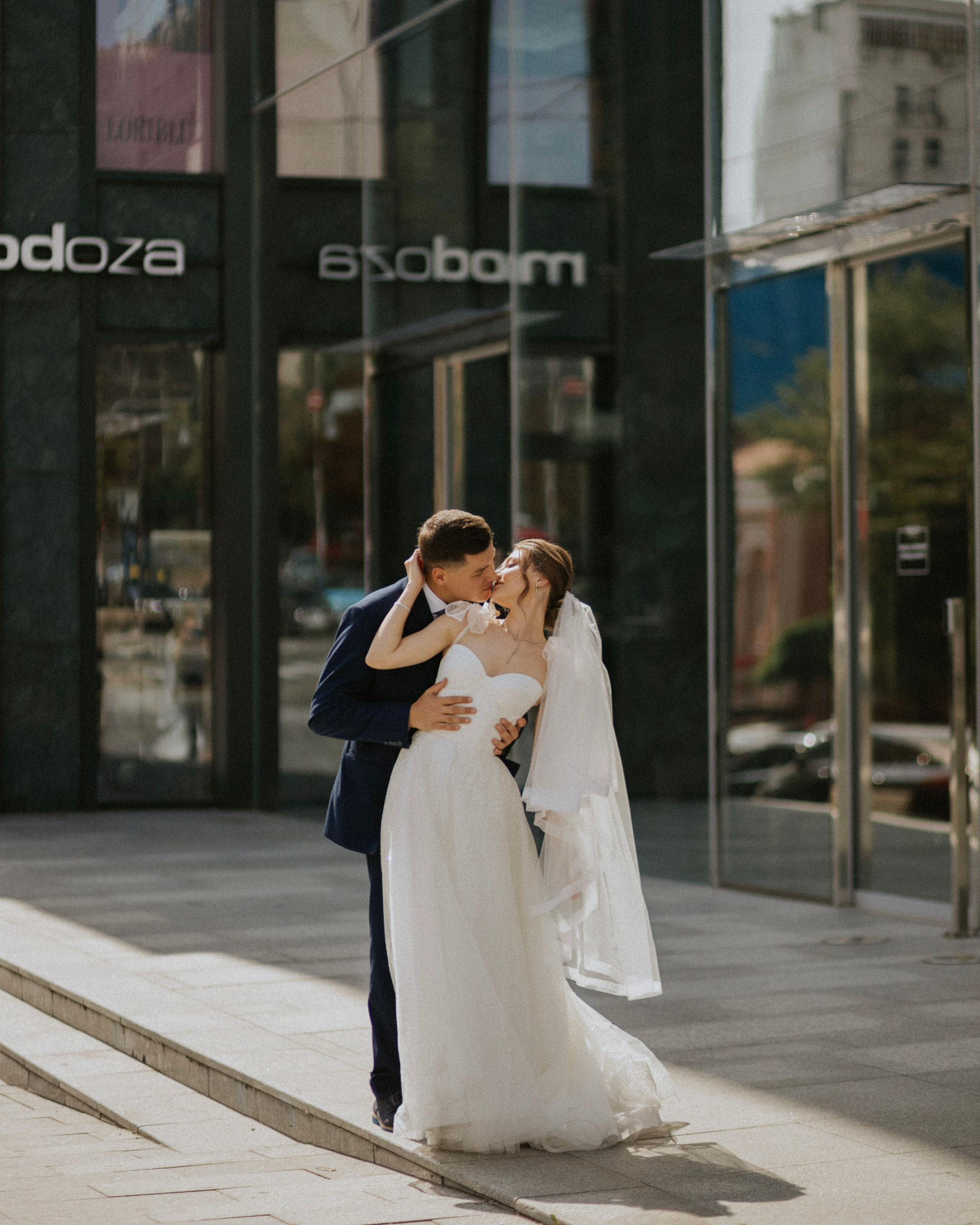 "Una coppia di sposi si bacia con gioia davanti a moderni edifici in vetro. La sposa, vestita con un fluente abito bianco, è in piedi sui gradini mentre lo sposo, che indossa un abito scuro, la solleva in un abbraccio affettuoso. Il momento è catturato in una vibrante ambientazione urbana”.