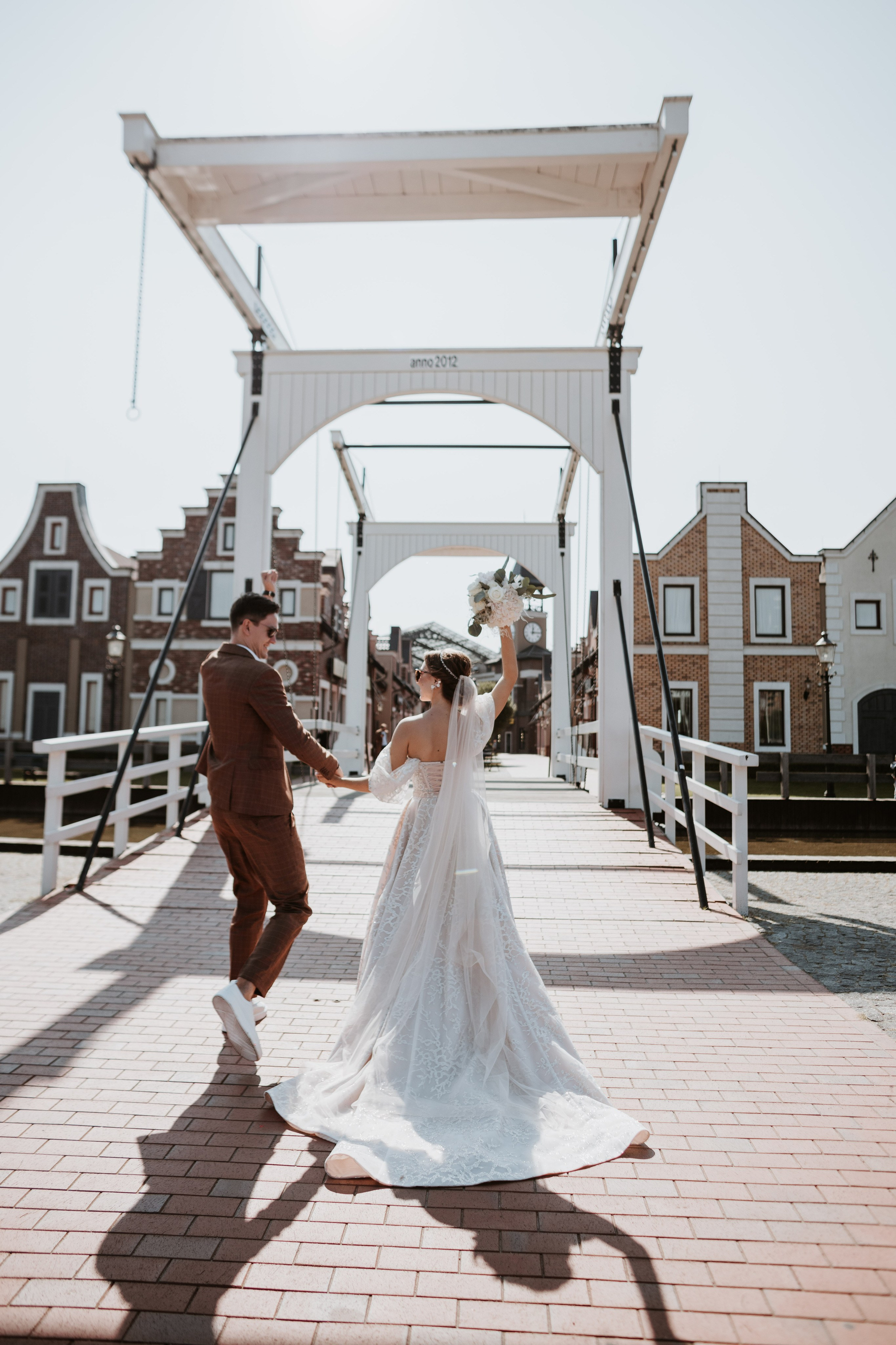 “Una sposa in abito di pizzo gira gioiosamente su un ponte, tenendo un bouquet in una mano. Il suo velo scorre dietro di lei mentre balla con il suo compagno, che indossa un abito marrone e scarpe da ginnastica bianche. Sono circondati da edifici caratteristici e il sole proietta una calda luce sulla scena”.