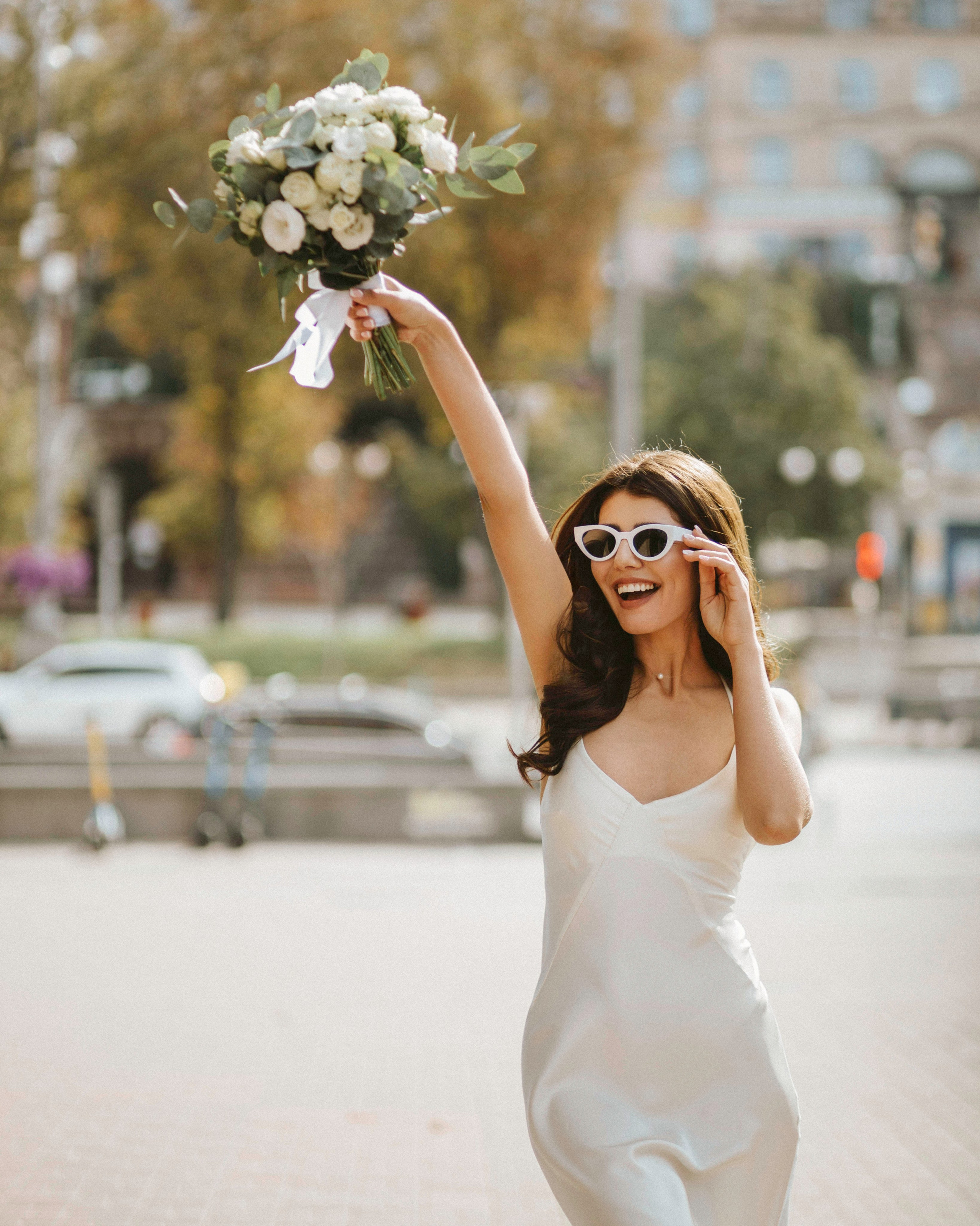 “Una sposa gioiosa in un elegante abito bianco che tiene in alto il suo bouquet e indossa eleganti occhiali da sole bianchi. Cammina all'aperto, sorridendo ampiamente alla luce del sole, con lo sfondo di una città vibrante”.