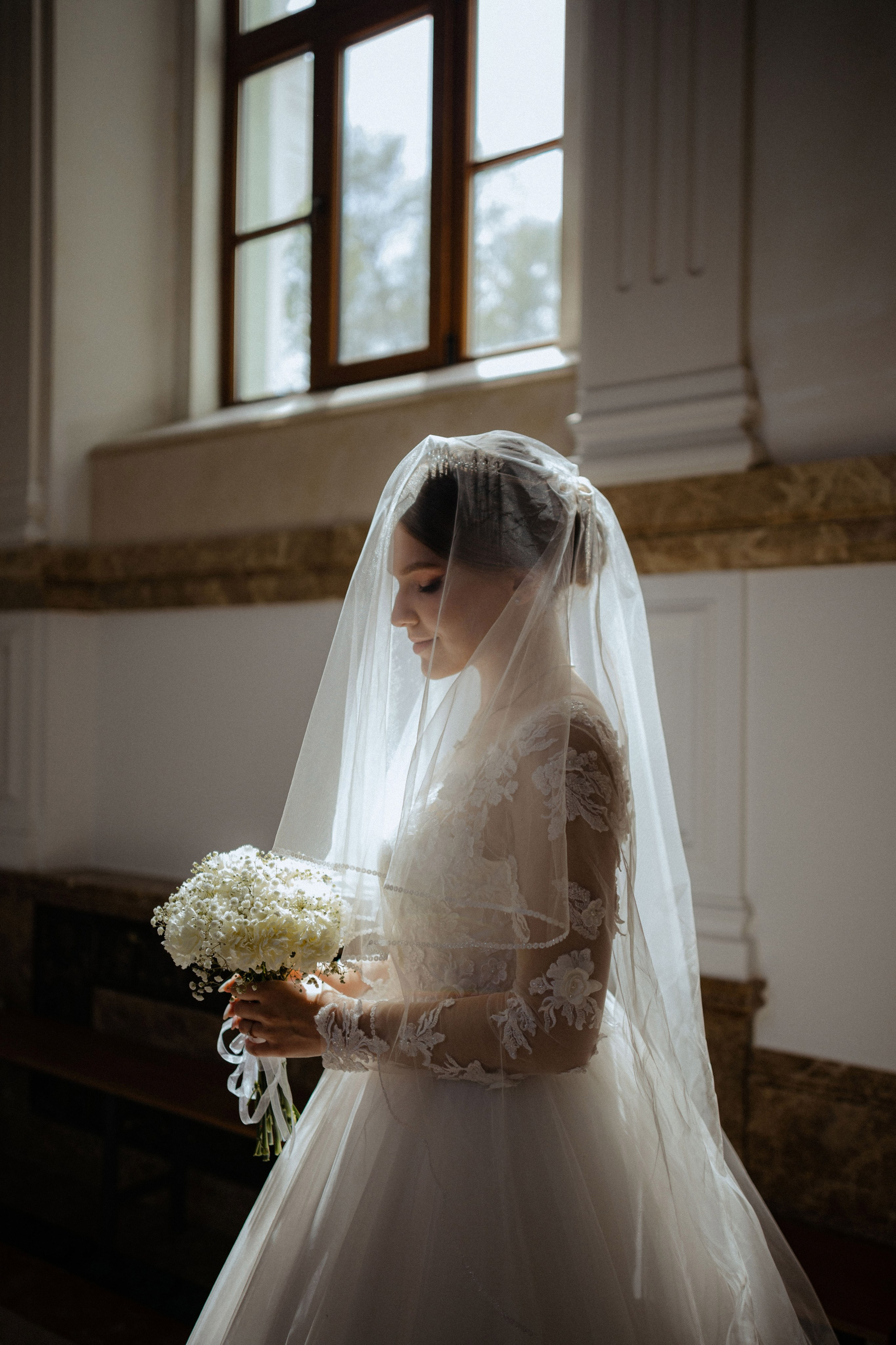 “Una sposa si erge con grazia, il suo volto è parzialmente illuminato dalla morbida luce del sole che filtra dalla finestra. Tiene in mano un bouquet di fiori bianchi mentre indossa un delicato abito da sposa in pizzo e un lungo velo fluente. Il momento sereno e intimo cattura la sua bellezza sullo sfondo morbido e neutro della stanza”.