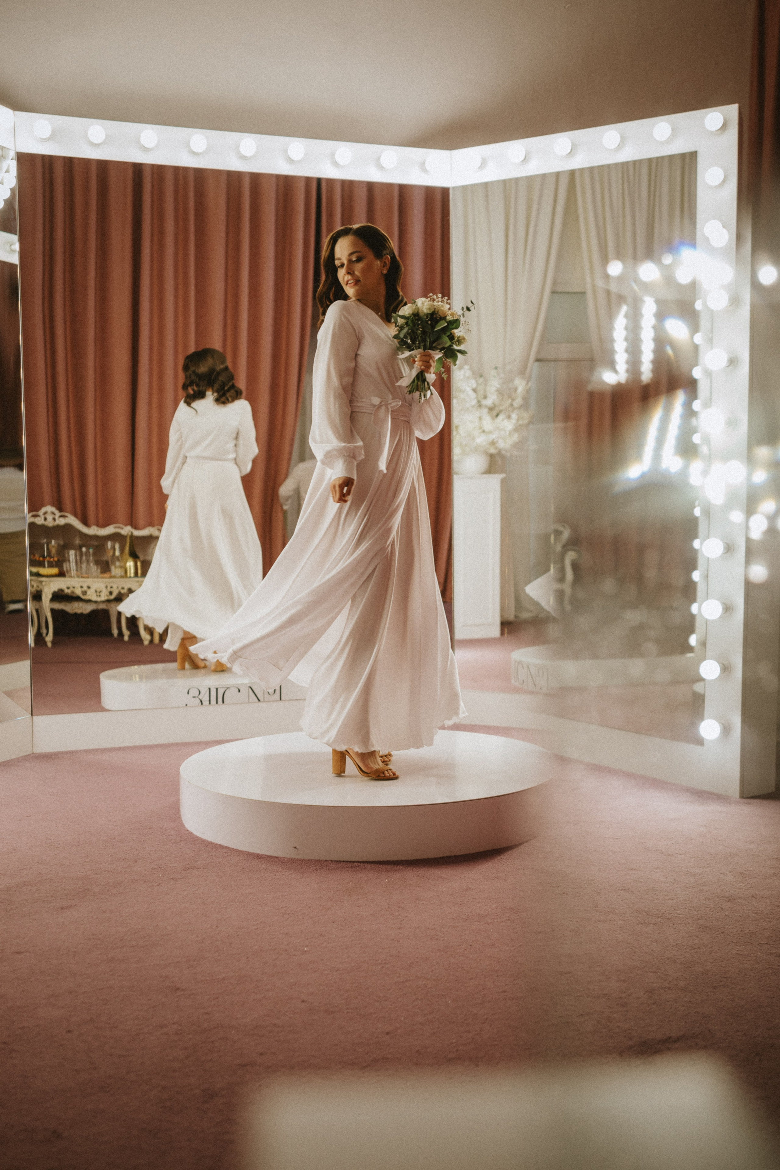 “Una sposa in una fluente vestaglia bianca volteggia con grazia su un piedistallo rotondo, tenendo in mano un bouquet di fiori freschi. L'ambientazione è un camerino glamour con grandi specchi luminosi che riflettono la sua immagine. Le luci rosa tenue e le decorazioni lussuose creano un'atmosfera calda e romantica, mentre lei sorride deliziata. La vestaglia della sposa scorre elegantemente mentre gira, catturando l'essenza dell'eleganza e della gioia nel suo giorno speciale”.