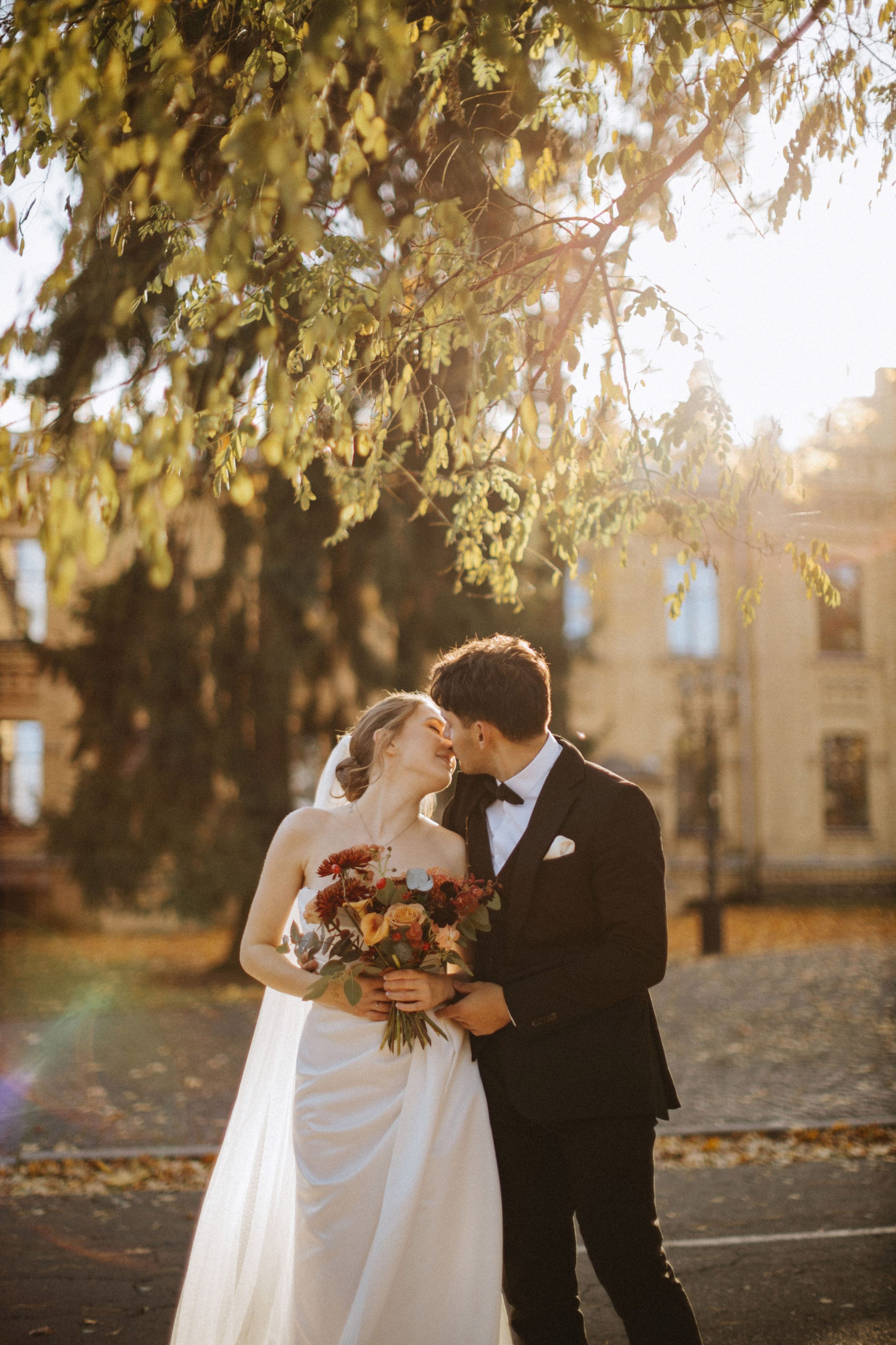 “Un momento romantico catturato tra la sposa e lo sposo mentre si scambiano un tenero bacio sotto la luce del sole. Circondata da vibranti foglie autunnali e dalla bellezza della natura, la sposa regge un bouquet di fiori dai toni caldi, creando un'ambientazione perfetta per un matrimonio autunnale.”