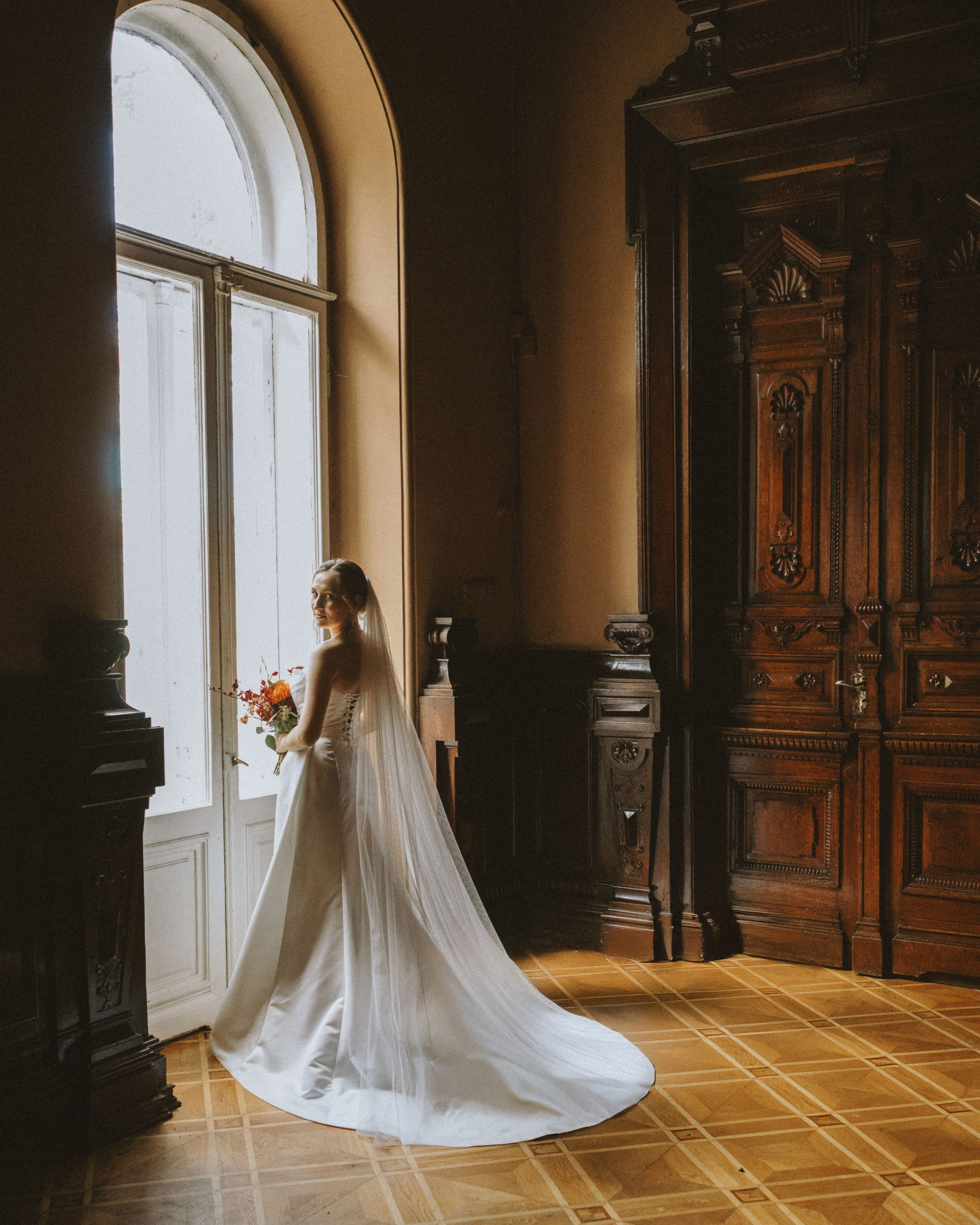 “Wedding photoshoot indoors. The bride stands by the window holding a bouquet, with her dress and veil flowing.”