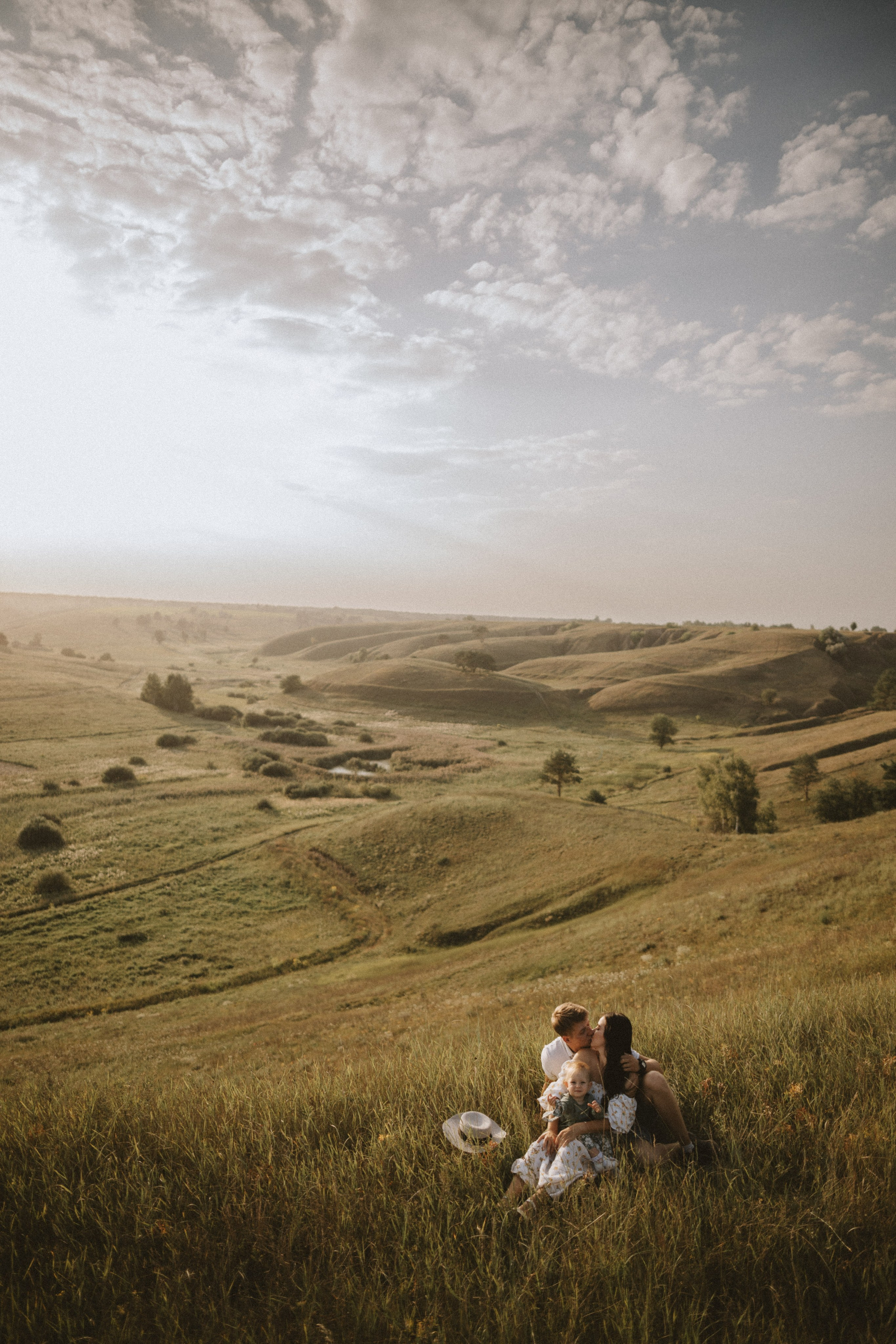“Una famiglia gioiosa che si gode un momento di pace nella natura, seduta su una collina erbosa. Il padre e la madre si scambiano un bacio mentre la madre tiene in grembo il figlio sorridente. Sullo sfondo si estende un vasto paesaggio di dolci colline, con un cielo limpido e una tenue luce solare che crea un caldo bagliore dorato”.