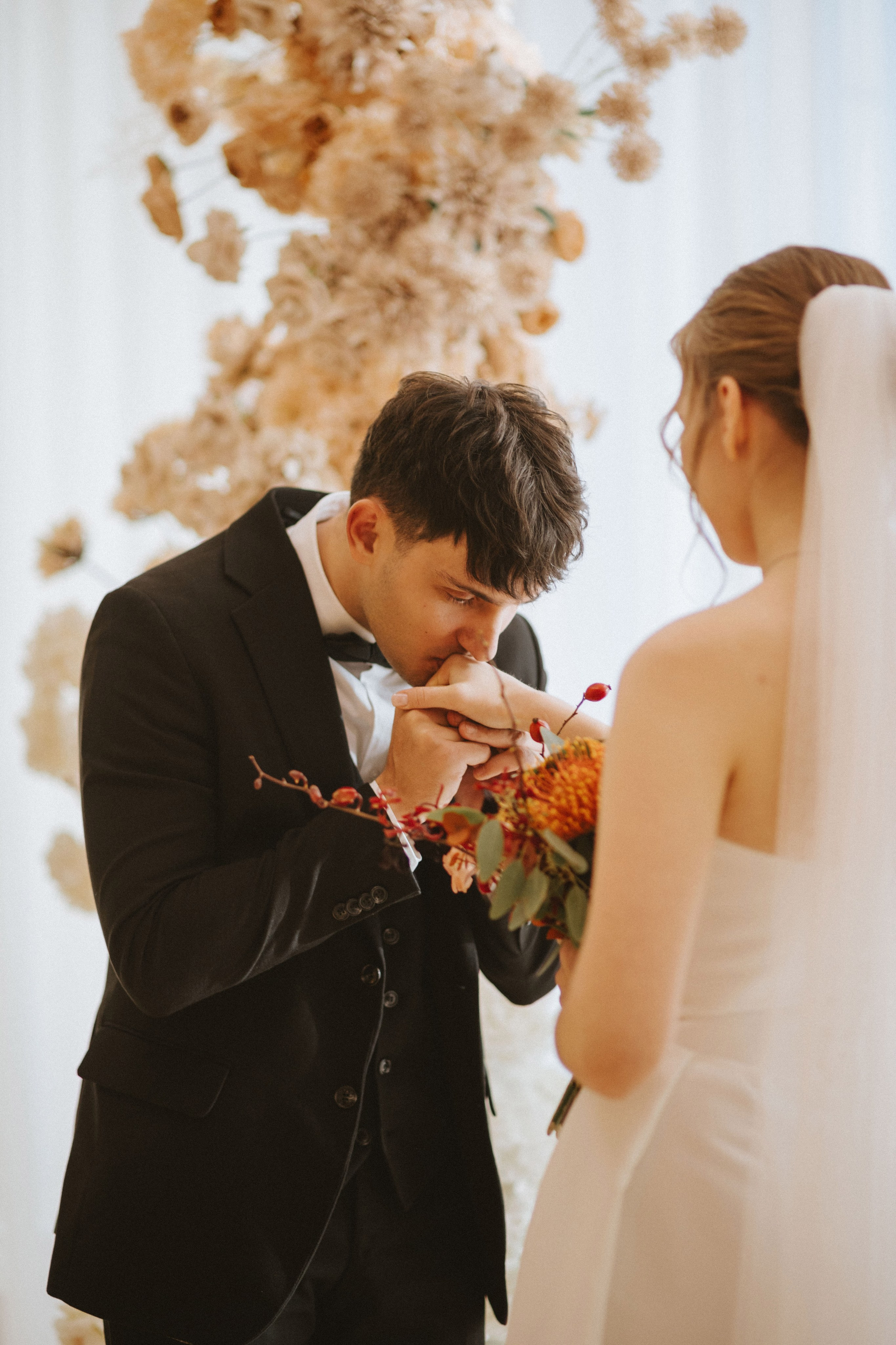 “Uno sposo, vestito con uno smoking nero, bacia delicatamente la mano della sua sposa, che si trova di fronte a lui con la schiena parzialmente visibile. Lei tiene in mano un bouquet di fiori e i due sono circondati da una composizione floreale dai toni neutri sullo sfondo. Il momento intimo è catturato in un'atmosfera calda e romantica”.
