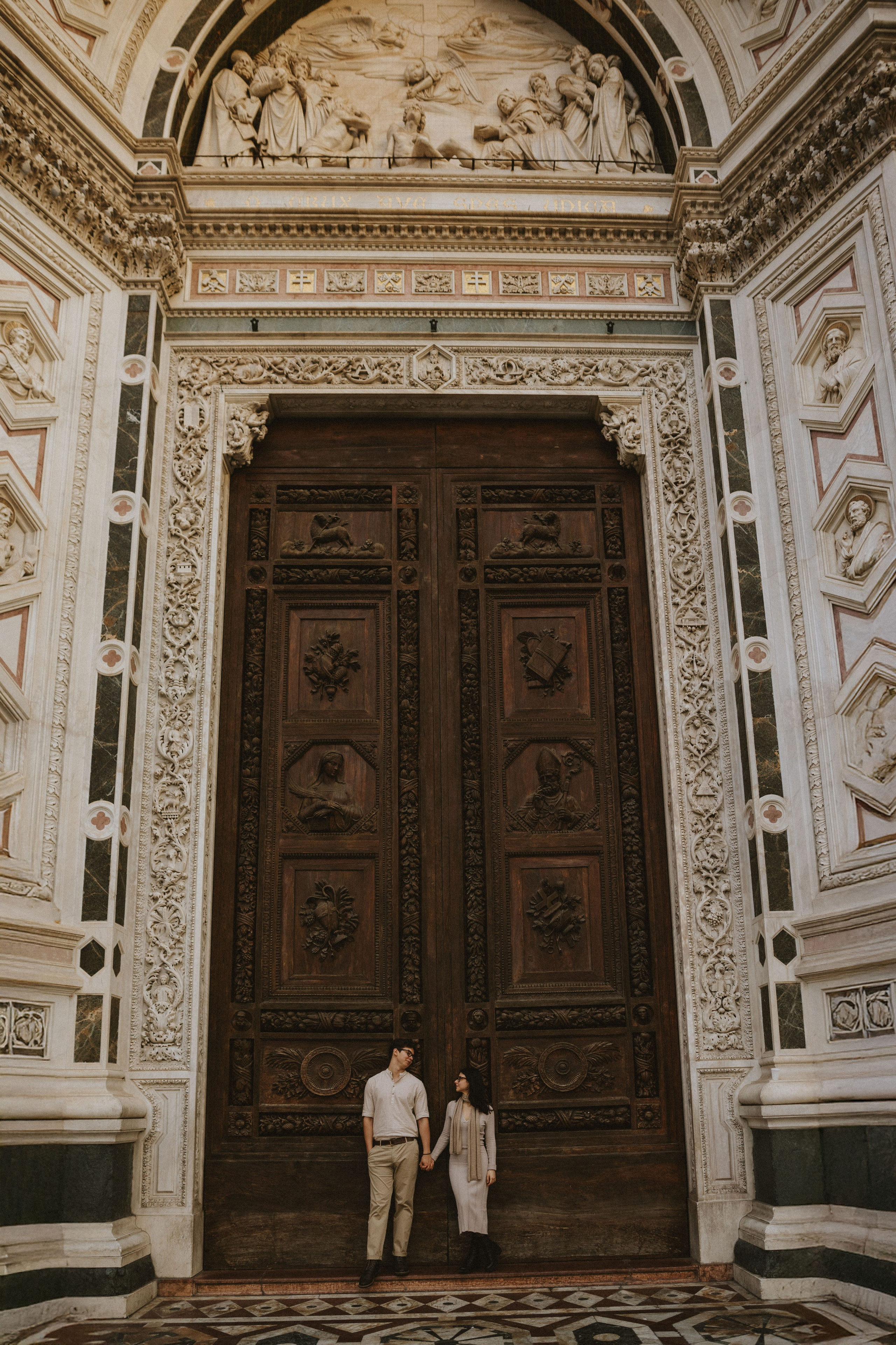 “Una coppia in piedi davanti a una porta di legno ornata con intricati intagli, che si tiene per mano e si guarda”.