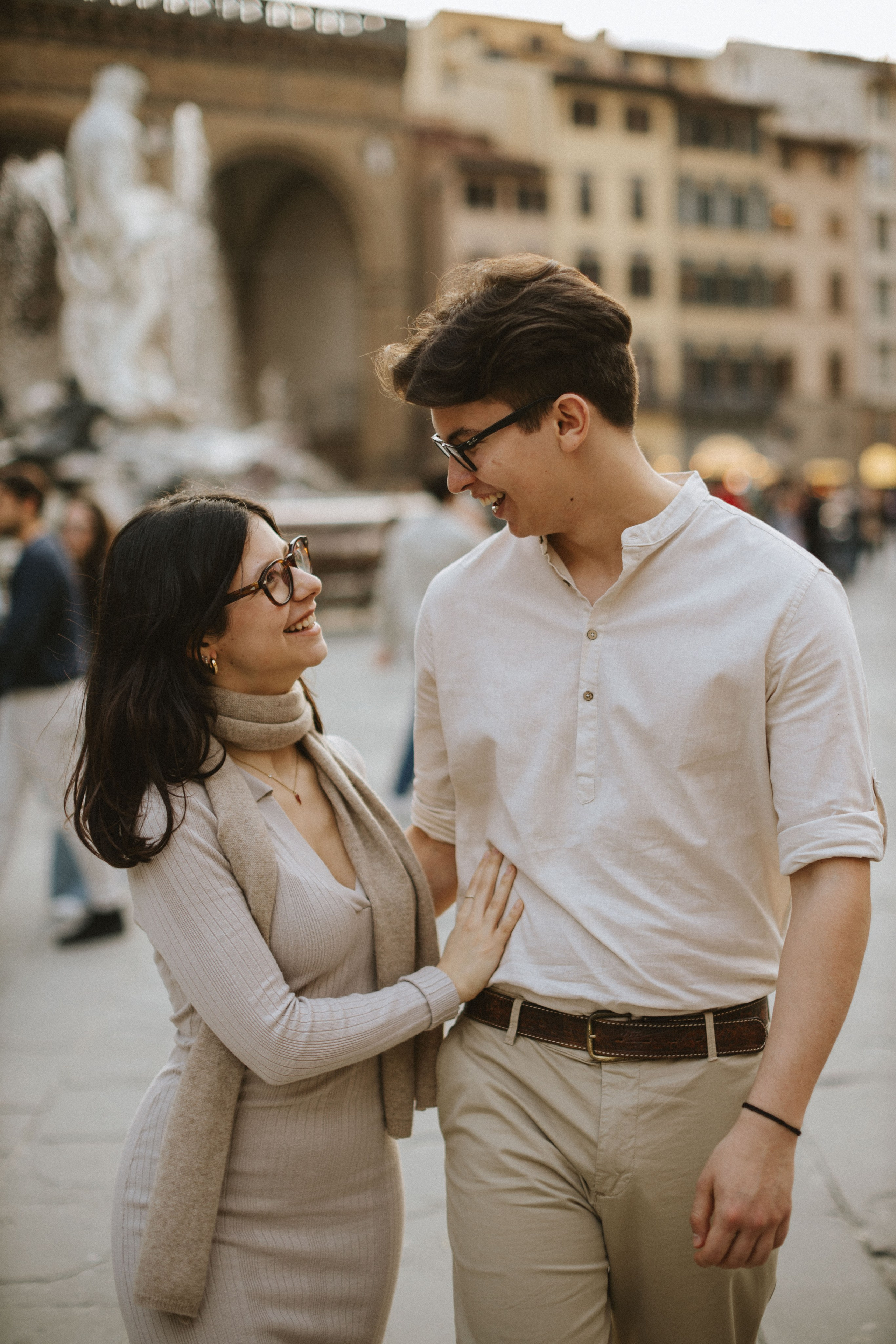 “Una coppia si sorride a vicenda, condividendo un momento di tenerezza in una piazza soleggiata con una fontana sullo sfondo”.