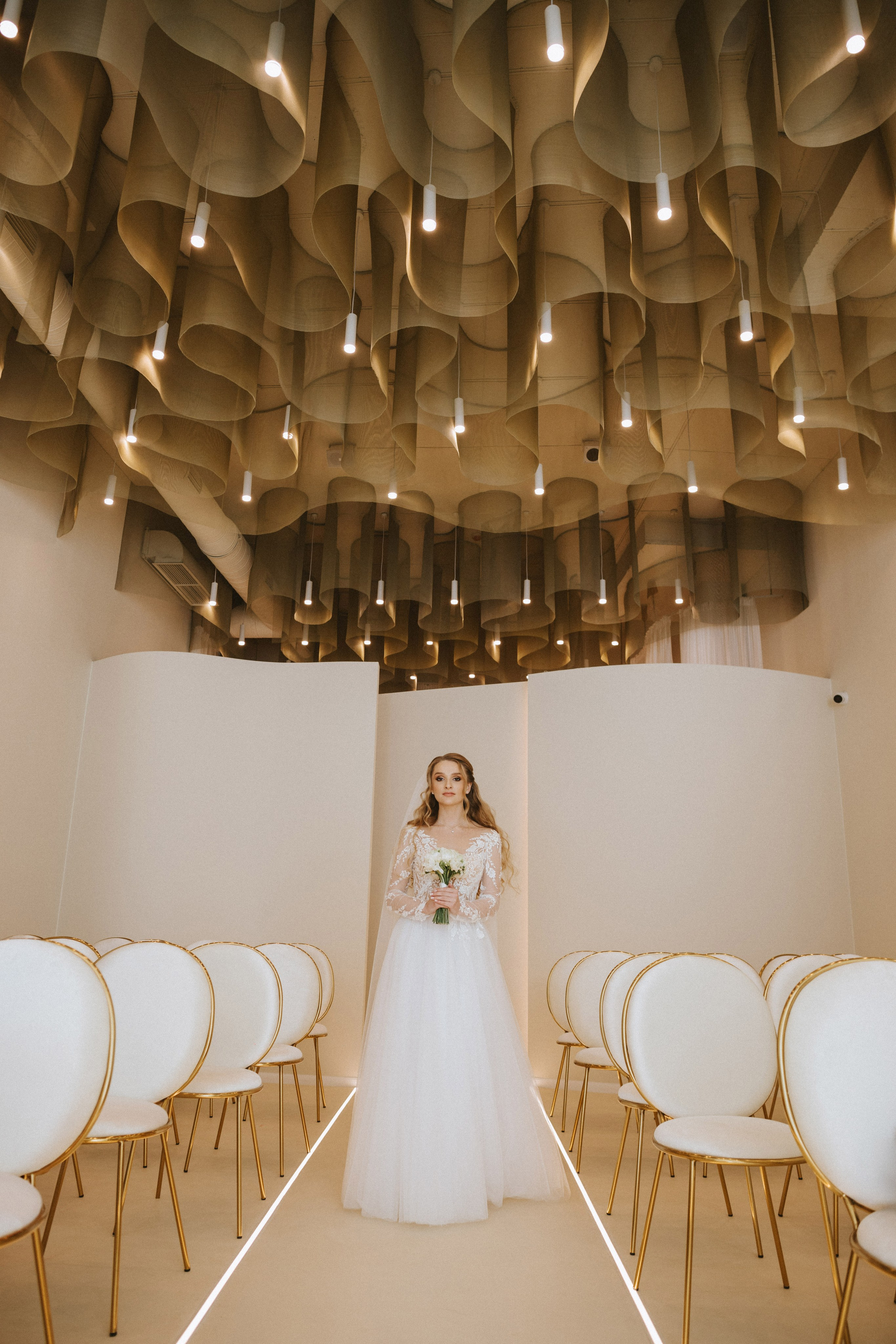 “Una sposa in un abito di pizzo a maniche lunghe si erge con grazia alla fine di una navata, tenendo in mano un bouquet di fiori bianchi. Il locale moderno presenta un soffitto elegante e curvilineo con luci a sospensione e sedie bianche eleganti disposte in file su entrambi i lati”.