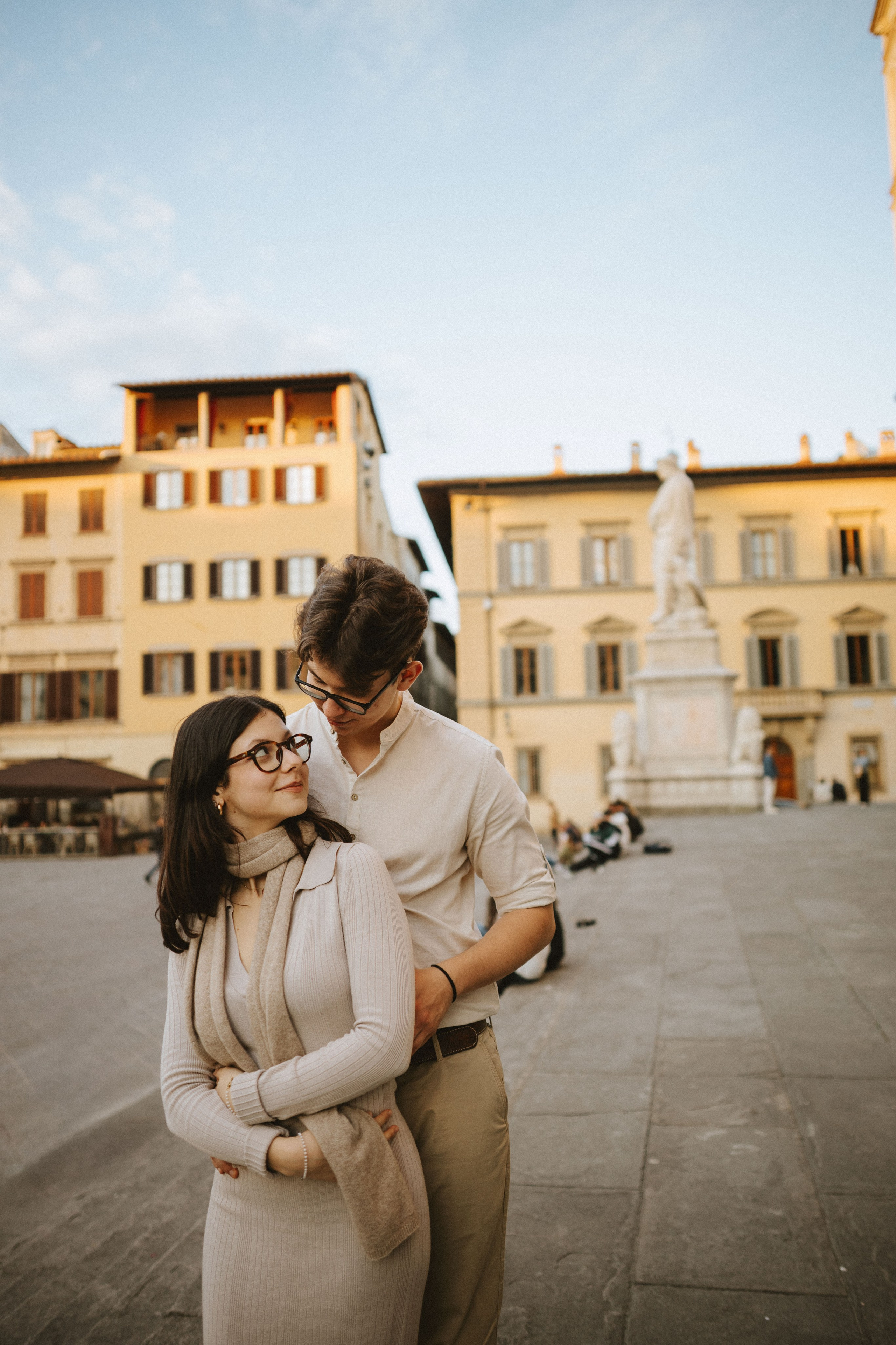 “Una coppia si abbraccia e si guarda in una piazza illuminata dal sole, con una statua sullo sfondo”.