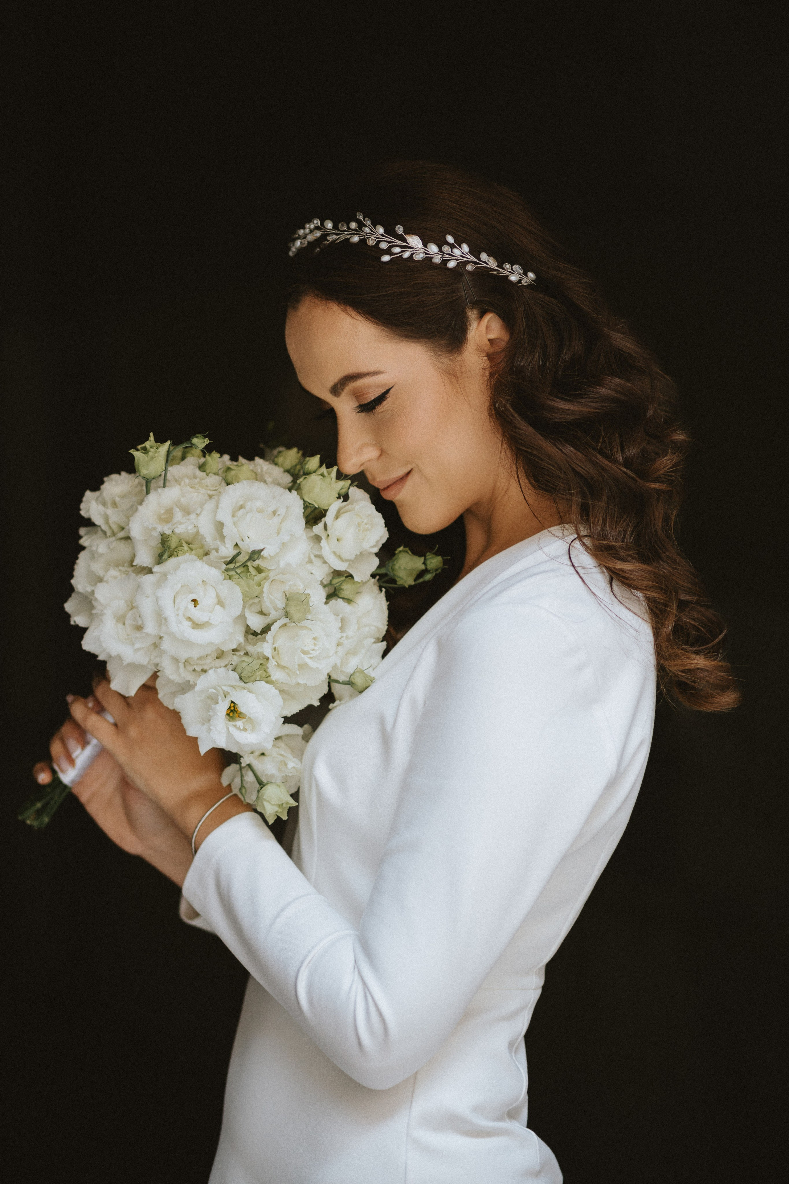 “Una sposa in un semplice ma elegante abito bianco, con in mano un bellissimo bouquet di rose bianche, guarda dolcemente i suoi fiori. I suoi capelli sono acconciati in morbidi riccioli e ornati da un delicato cerchietto di perle. Il contrasto dello sfondo scuro mette in risalto la sua espressione serena e aggraziata”.