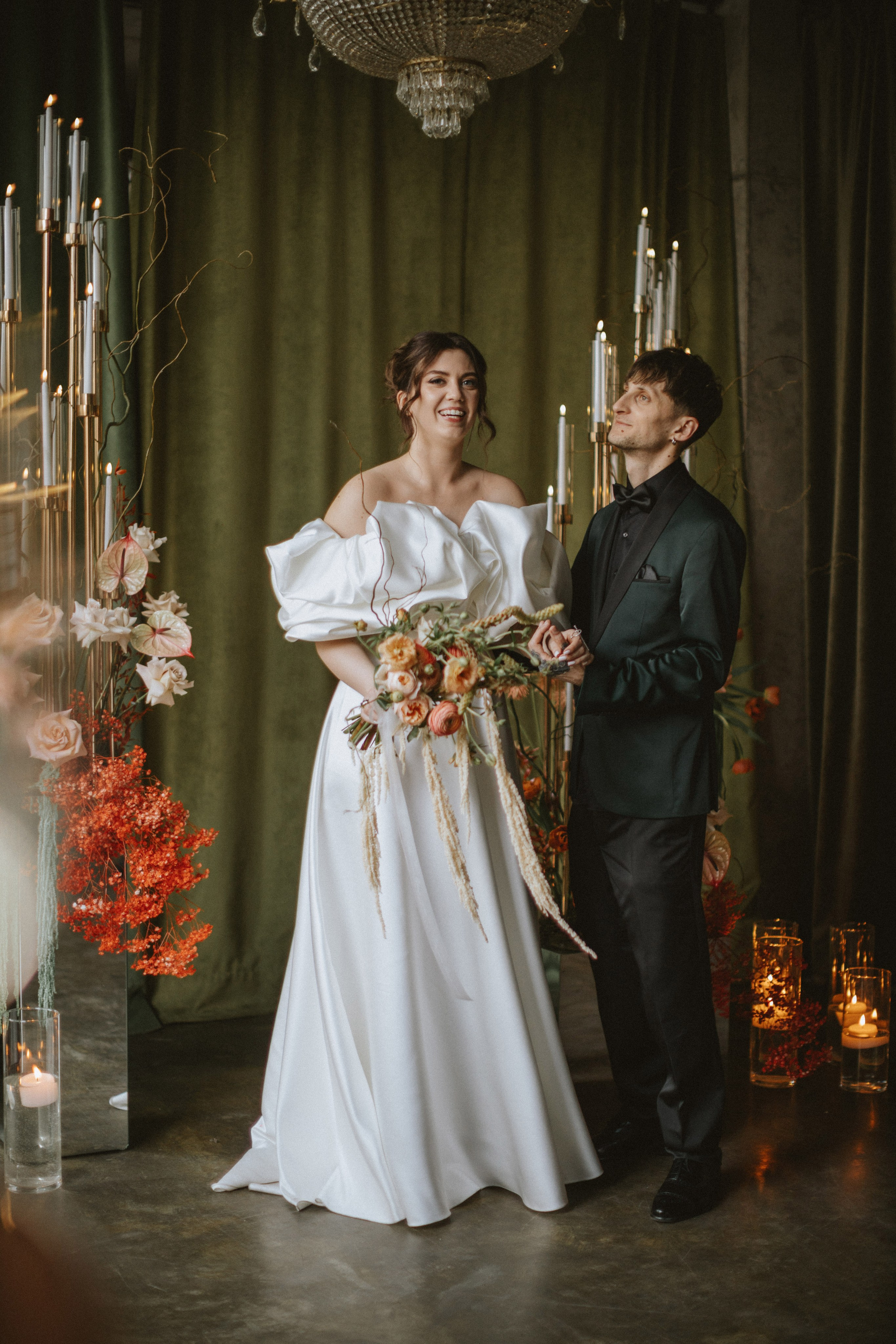 “Una sposa e uno sposo in piedi insieme in un ambiente romantico, circondati da candele e composizioni floreali. La sposa sorride gioiosa tenendo in mano un bouquet di fiori, indossando un voluminoso abito bianco con un drammatico design off-shoulder. Lo sposo, in smoking verde con papillon nero, la guarda con affetto. La scena è immersa in una luce calda e morbida”.