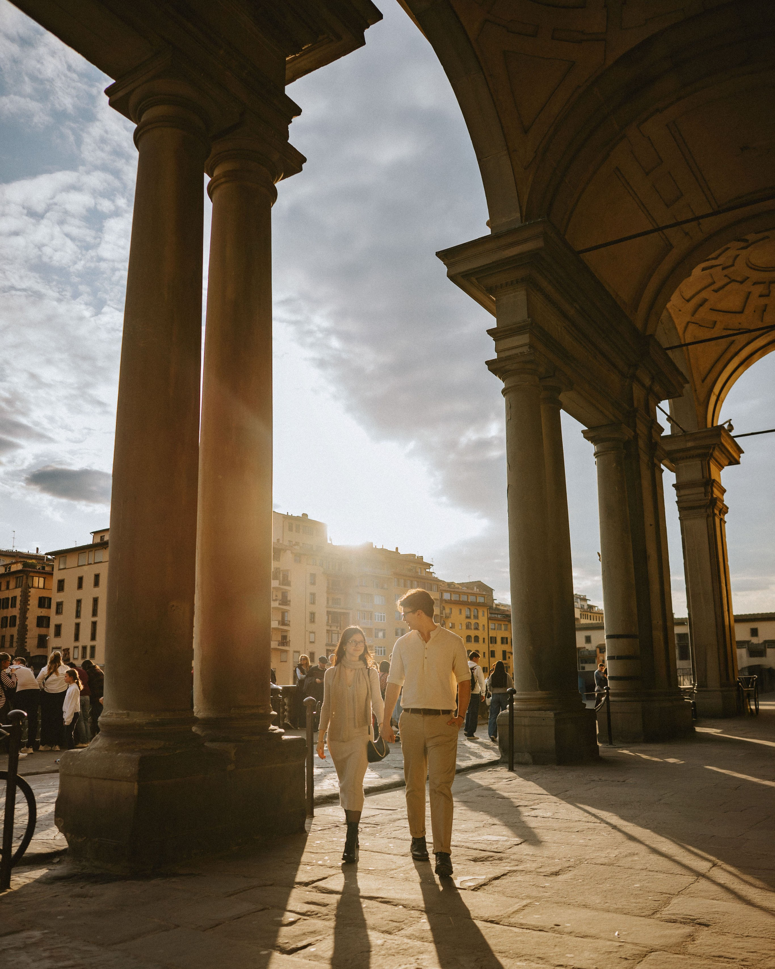 “A couple strolling under arches with a sunset in the background, in the historic city center.”