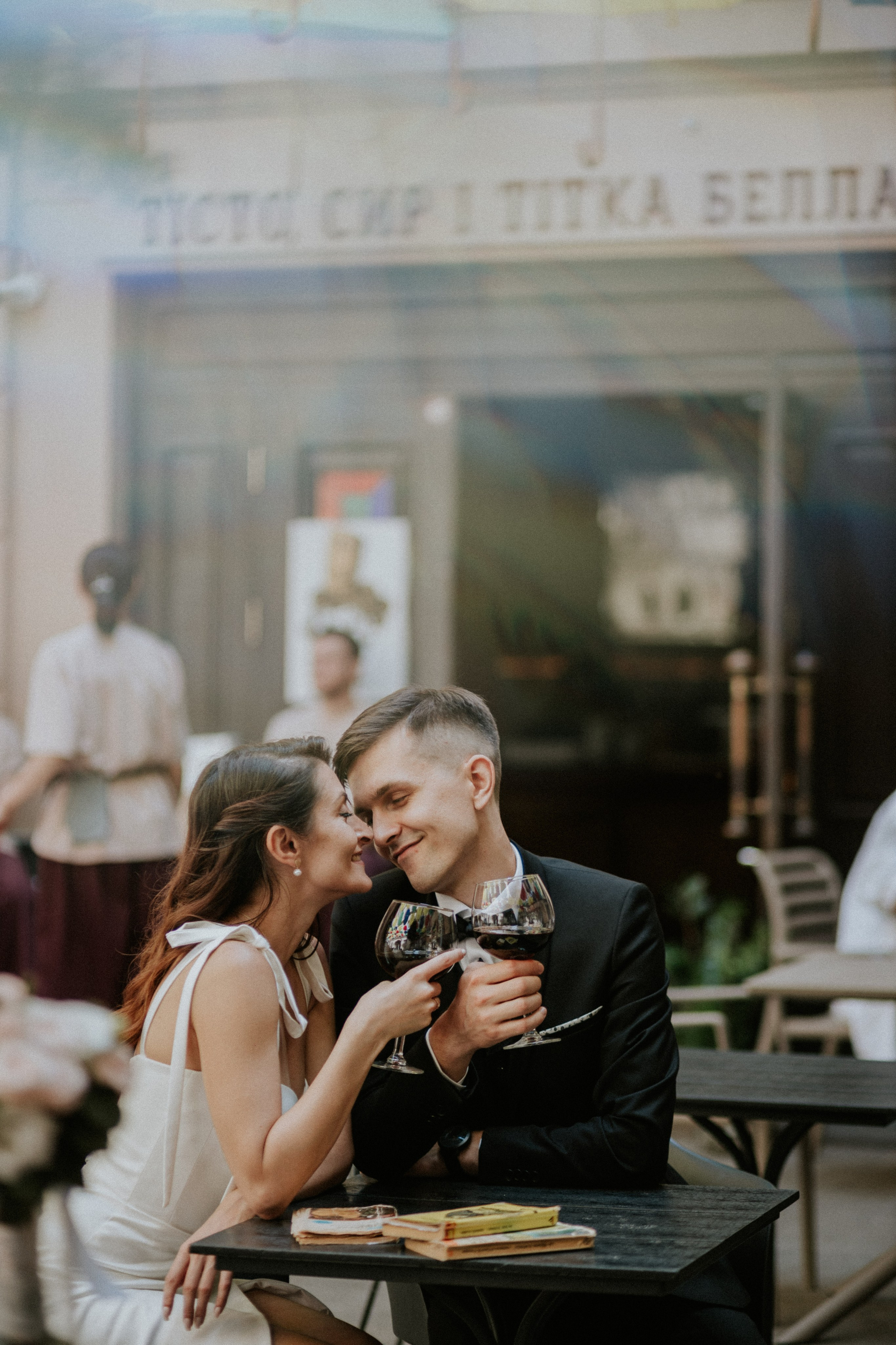 “Una coppia di sposi che condivide un momento romantico in un caffè all'aperto, sorridendosi amorevolmente mentre tiene in mano dei bicchieri di vino rosso. La donna indossa un elegante abito da sposa bianco con un dettaglio di fiocco sulla schiena, mentre l'uomo è vestito con un abito nero e un papillon. Lo sfondo è caratterizzato da un'insegna del ristorante sfocata e da un'atmosfera rilassata con altre persone sullo sfondo”.