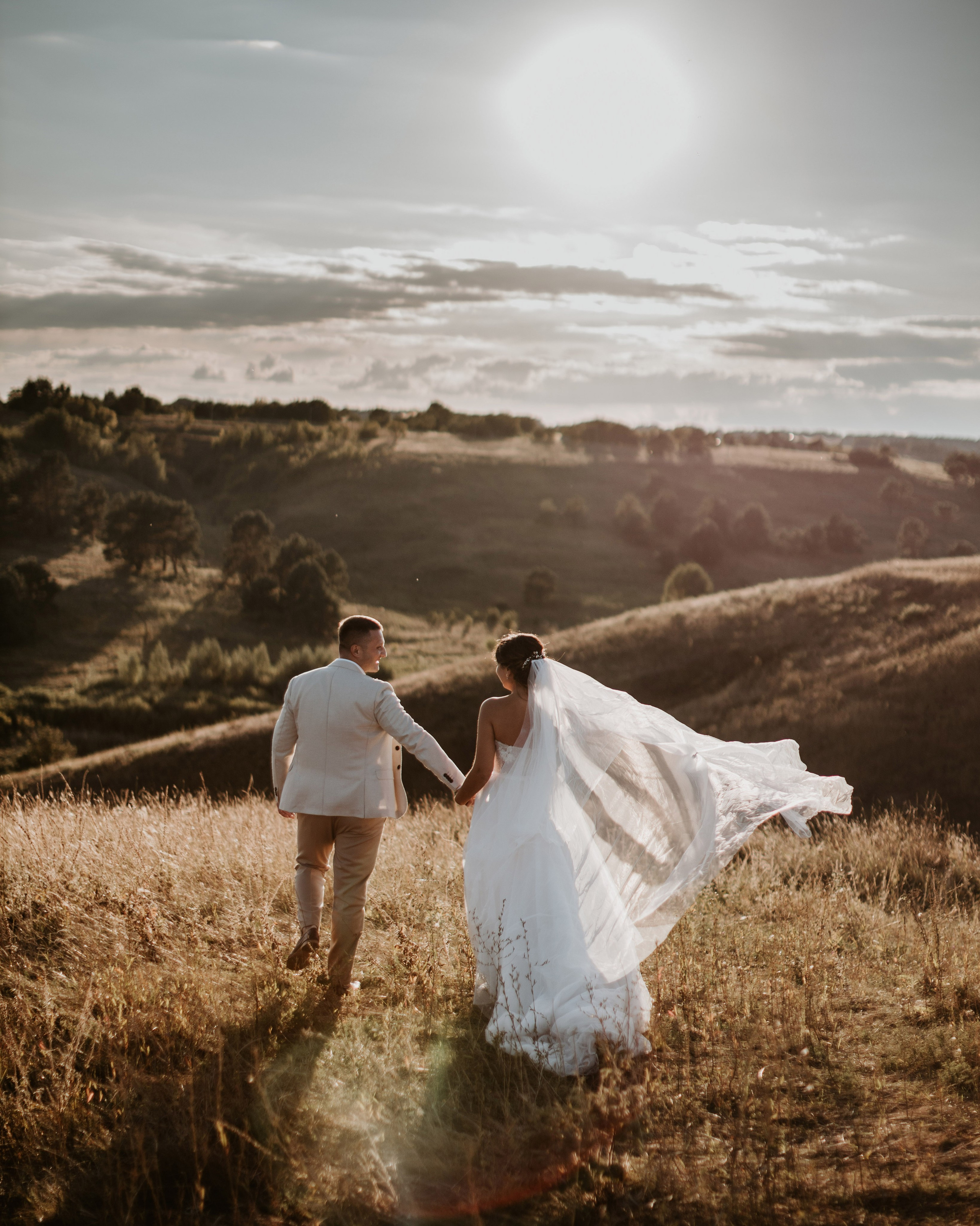“Wedding photoshoot in nature. The bride and groom hold hands, walking on a green hill with the wind blowing the bride’s veil.”