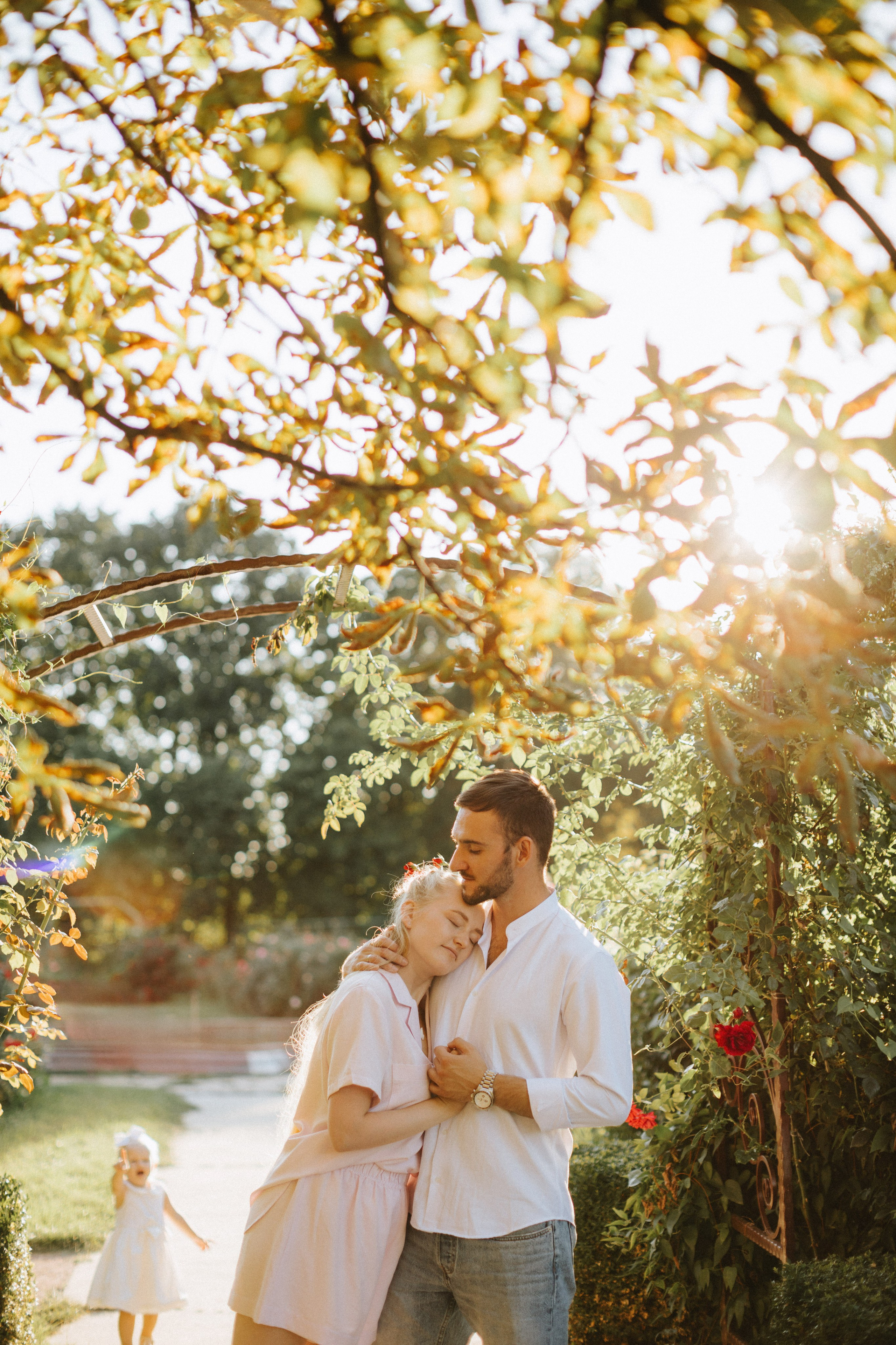 “Una coppia abbracciata in un giardino, con la donna che appoggia la testa sul petto dell'uomo. Sullo sfondo, il loro bambino in abito bianco cammina verso di loro. La scena è illuminata da una luce solare morbida e dorata che filtra attraverso le foglie”.