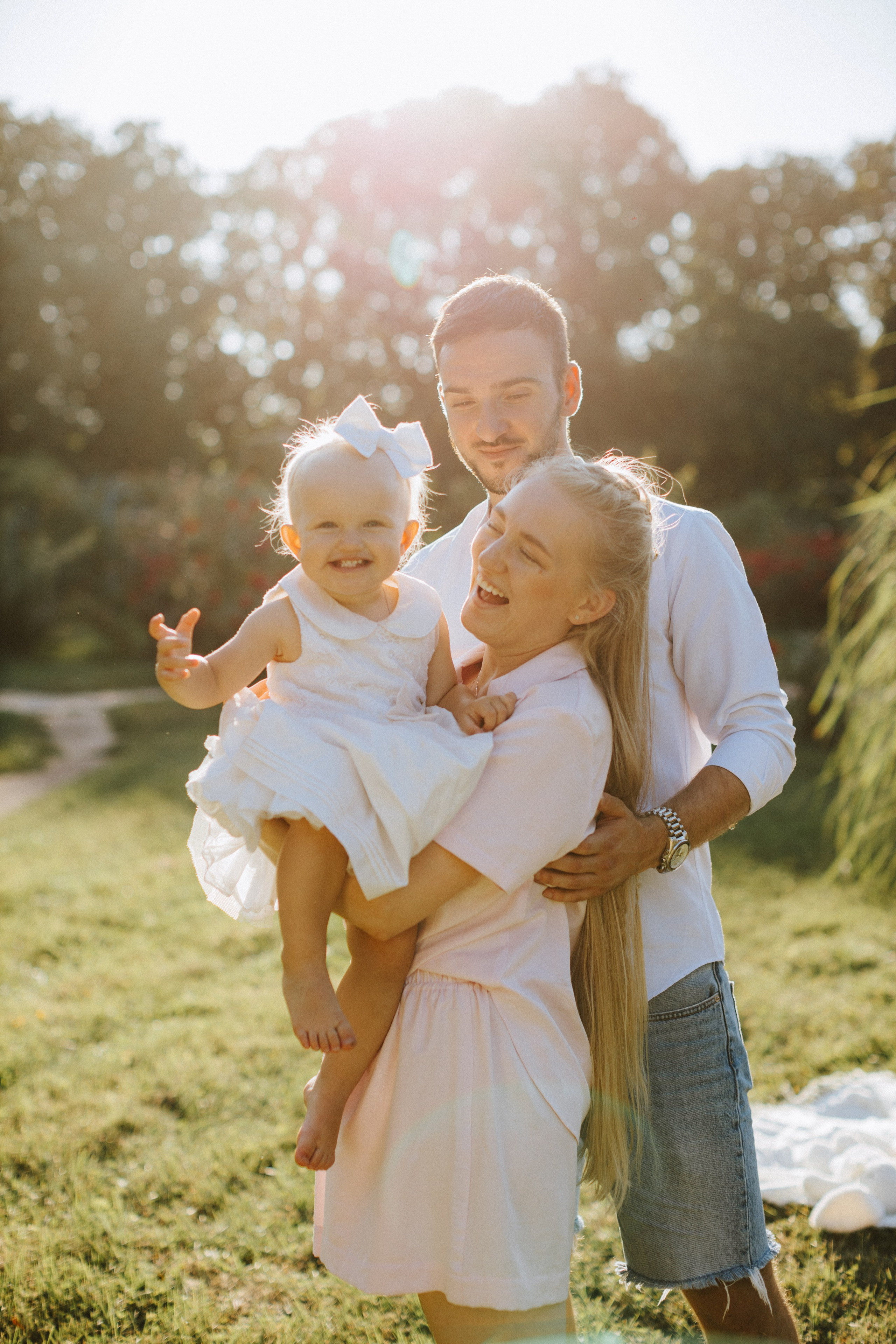 Una famiglia felice che si gode una giornata di sole nel parco. La madre tiene in braccio la figlia sorridente, mentre il padre è accanto a loro e sorride calorosamente. La bambina indossa un vestito bianco con un grande fiocco e irradia gioia. La luce del sole aggiunge una calda luminosità alla scena, circondata dal verde dell'erba e dalla natura sullo sfondo”.