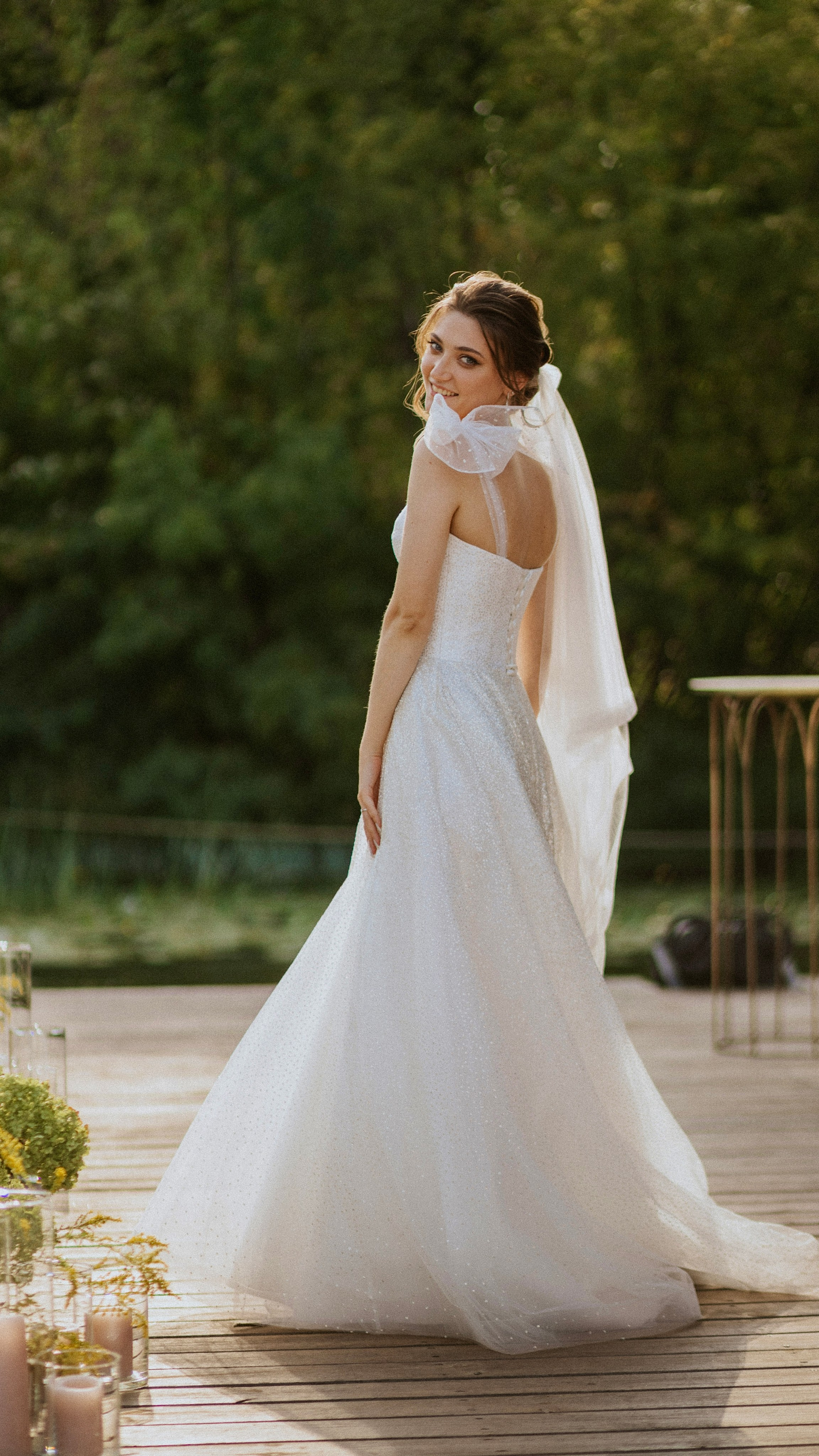 “Una sposa, che indossa uno splendido abito bianco con delicati scintillii, si gira con grazia per guardarsi alle spalle. L'abito scorre magnificamente dietro di lei mentre cammina lungo una piattaforma di legno, con una morbida vegetazione che circonda la scena. La luce filtra attraverso il velo e crea un bagliore caldo, aggiungendo un'atmosfera serena e magica al momento”.