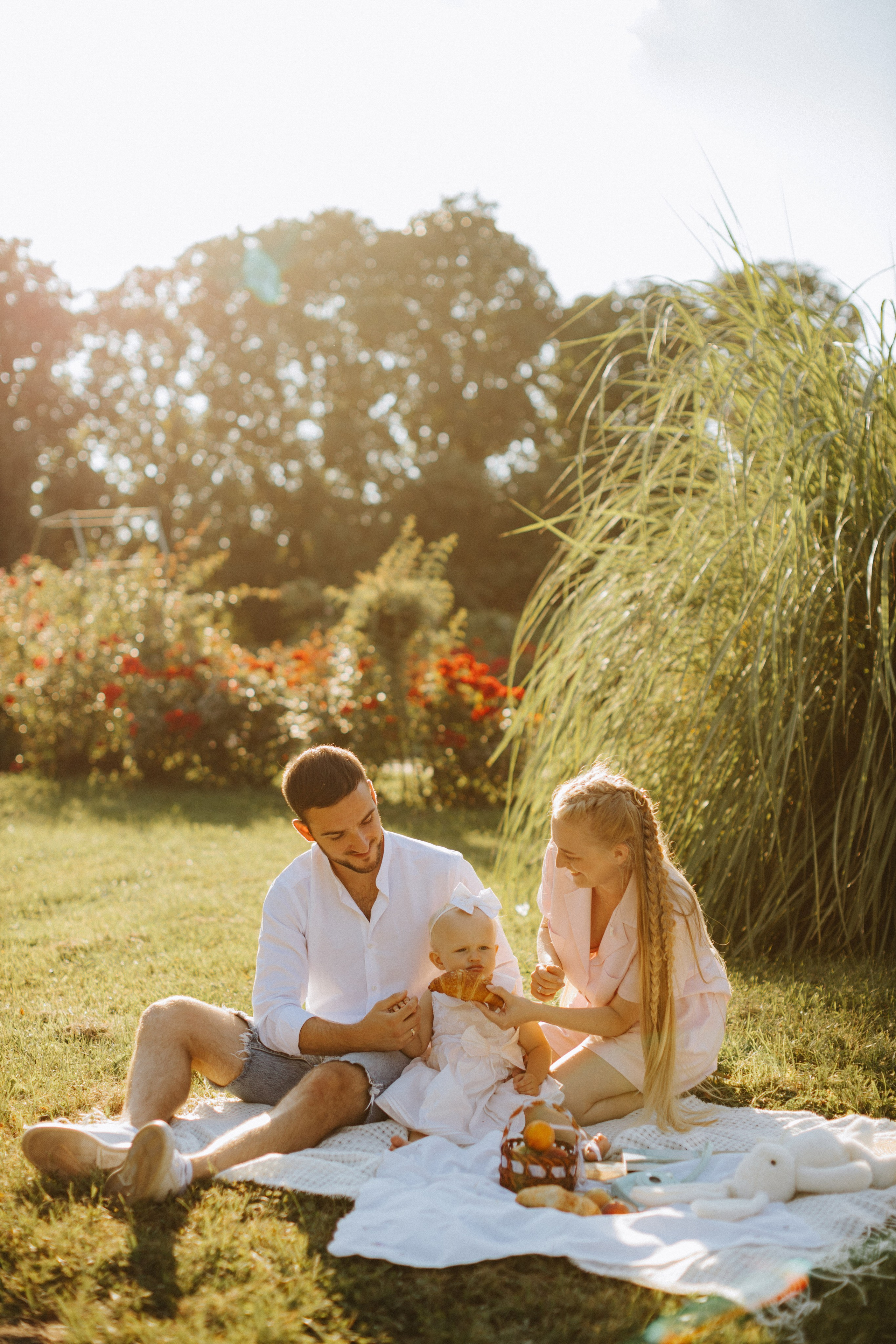 “Una famiglia di tre persone si gode un picnic all'aperto. Il padre tiene in braccio la figlia che mangia un croissant, mentre la madre sorride loro. Lo sfondo è caratterizzato da una vegetazione lussureggiante e da fiori vivaci, che creano un'atmosfera calda e gioiosa”.