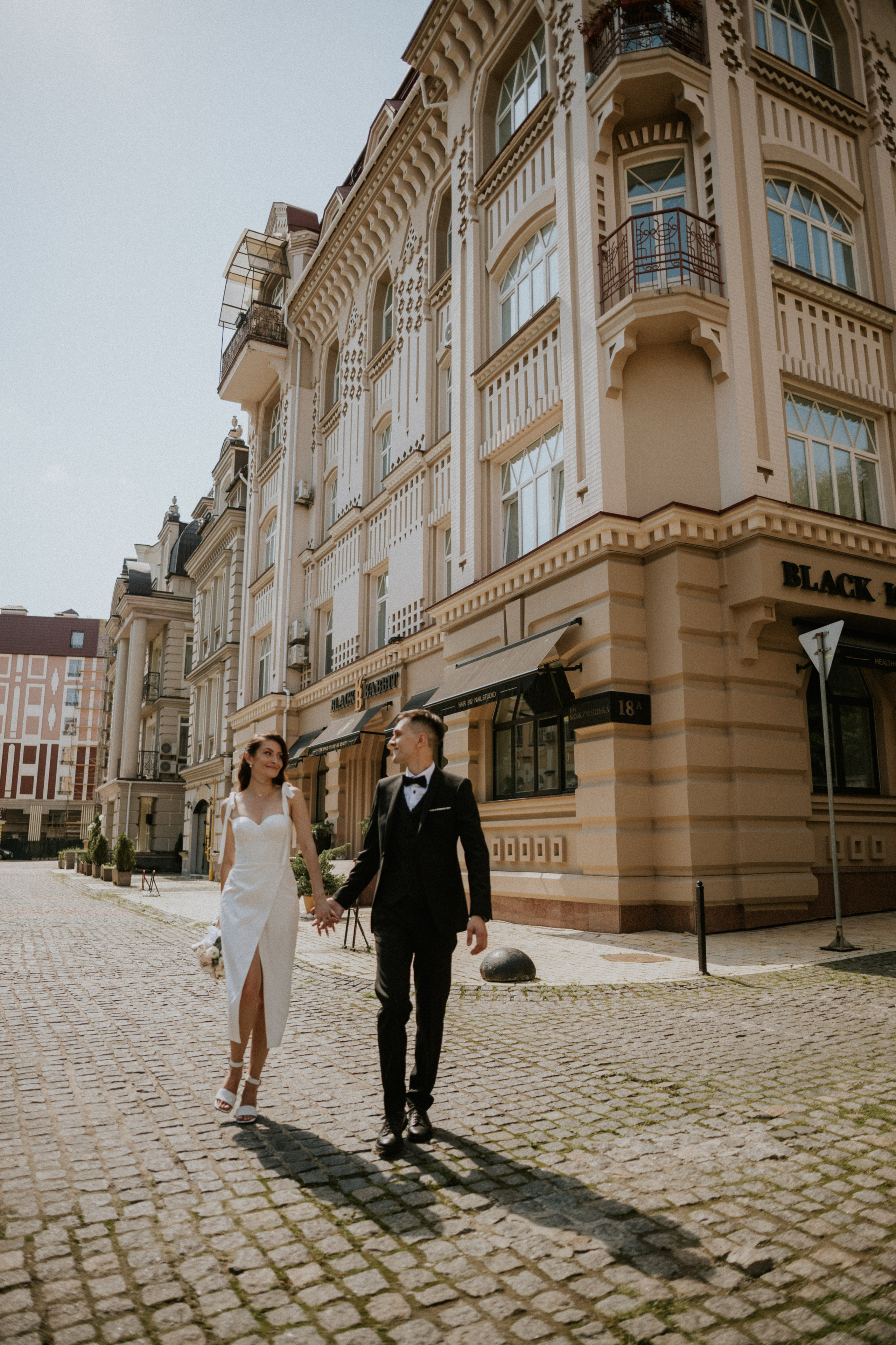 “Una coppia di sposi passeggia in un'affascinante strada cittadina, mano nella mano. La sposa, che indossa un elegante abito bianco con spacco alto, cammina con sicurezza accanto allo sposo, vestito con uno smoking classico. I due condividono un momento di gioia e di intesa, mentre gli eleganti edifici sullo sfondo aggiungono un'atmosfera sofisticata alla scena. La coppia si sorride a vicenda, irradiando felicità contro la calda luce del sole”.