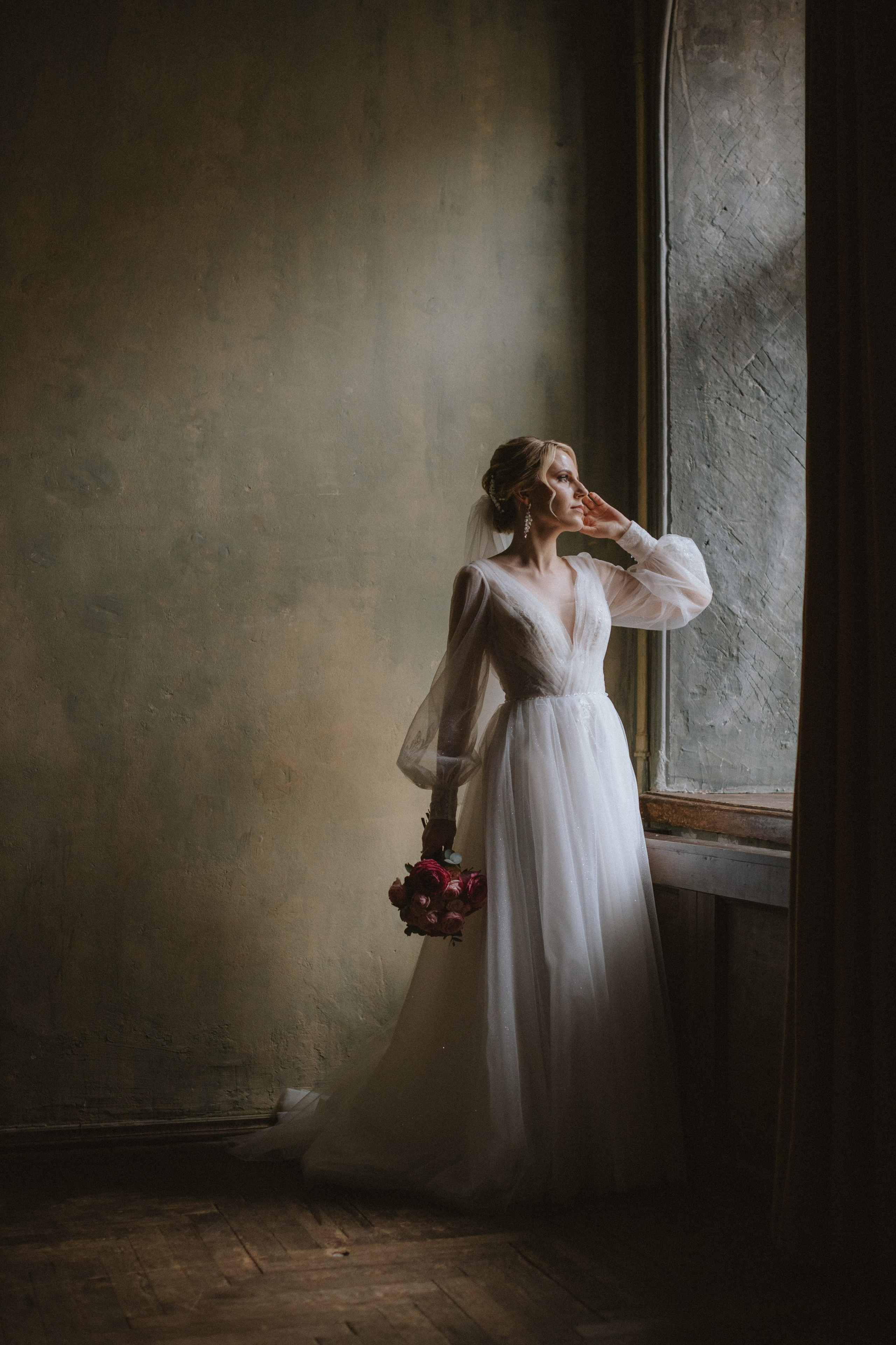“Una sposa in un abito leggero e trasparente con maniche lunghe si trova vicino a una finestra, con in mano un bouquet di fiori rosa. Guarda pensierosa fuori dalla finestra, con un'espressione serena e contemplativa, mentre la luce naturale soffusa illumina i suoi lineamenti delicati”.