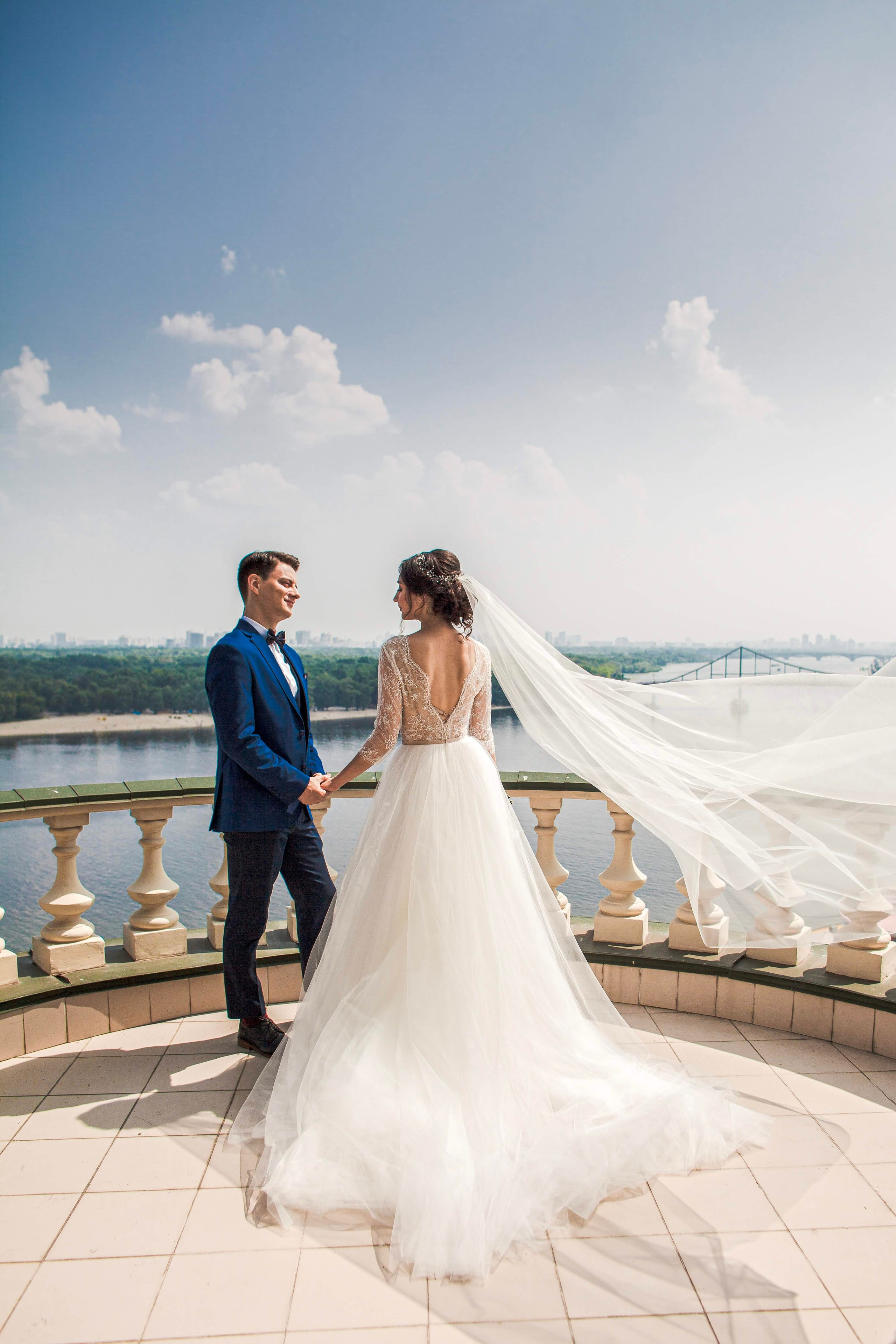 “Una sposa e uno sposo in piedi su un balcone con una splendida vista sul fiume sullo sfondo. La sposa indossa un fluente abito bianco con un lungo velo che viene delicatamente mosso dal vento, mentre lo sposo è in abito blu con papillon. Si tengono per mano e si sorridono a vicenda, godendosi il momento sotto un cielo limpido”.