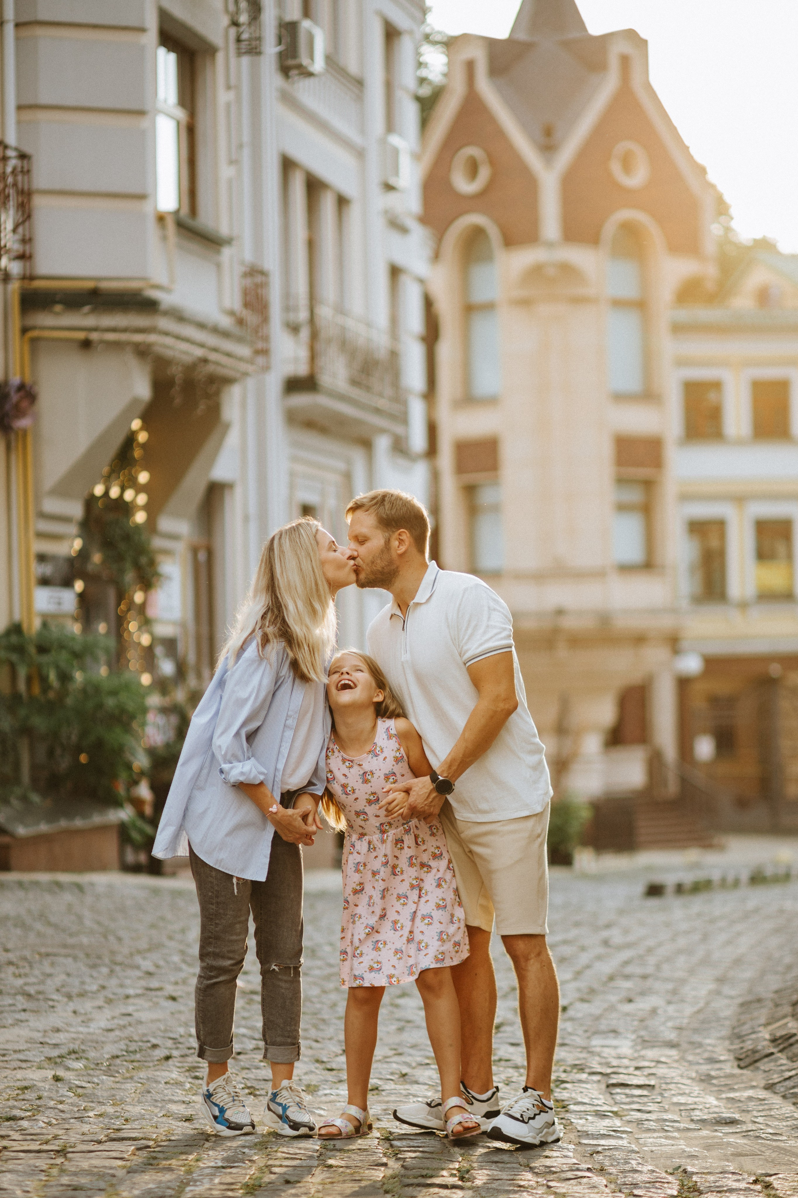 “Un gioioso momento di famiglia in città: padre e madre si scambiano un bacio, mentre la loro giovane figlia ride di gioia. La luce dorata del sole serale aggiunge calore alla scena, evidenziando il legame felice e affettuoso tra i due. Sono in piedi su una strada acciottolata con affascinanti edifici cittadini sullo sfondo”.