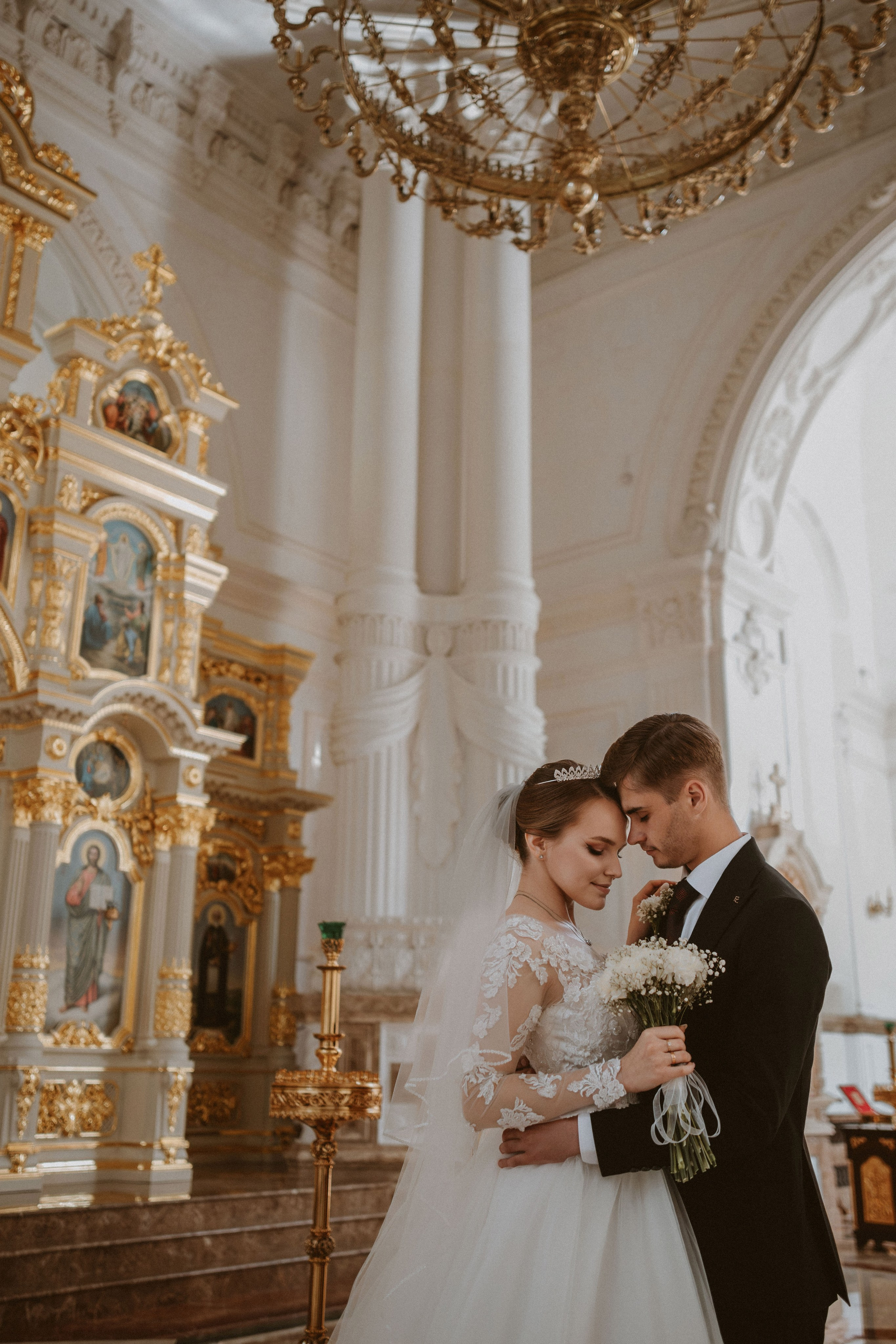 “Una coppia condivide un momento di tenerezza in una splendida chiesa, circondata da grandi icone dorate e da uno splendido lampadario. La sposa, che indossa un delicato abito da sposa in pizzo e una tiara, tiene in mano un bouquet di fiori bianchi, mentre lo sposo, vestito con un abito classico, la abbraccia dolcemente. Le loro fronti si toccano in un gesto intimo e affettuoso”.