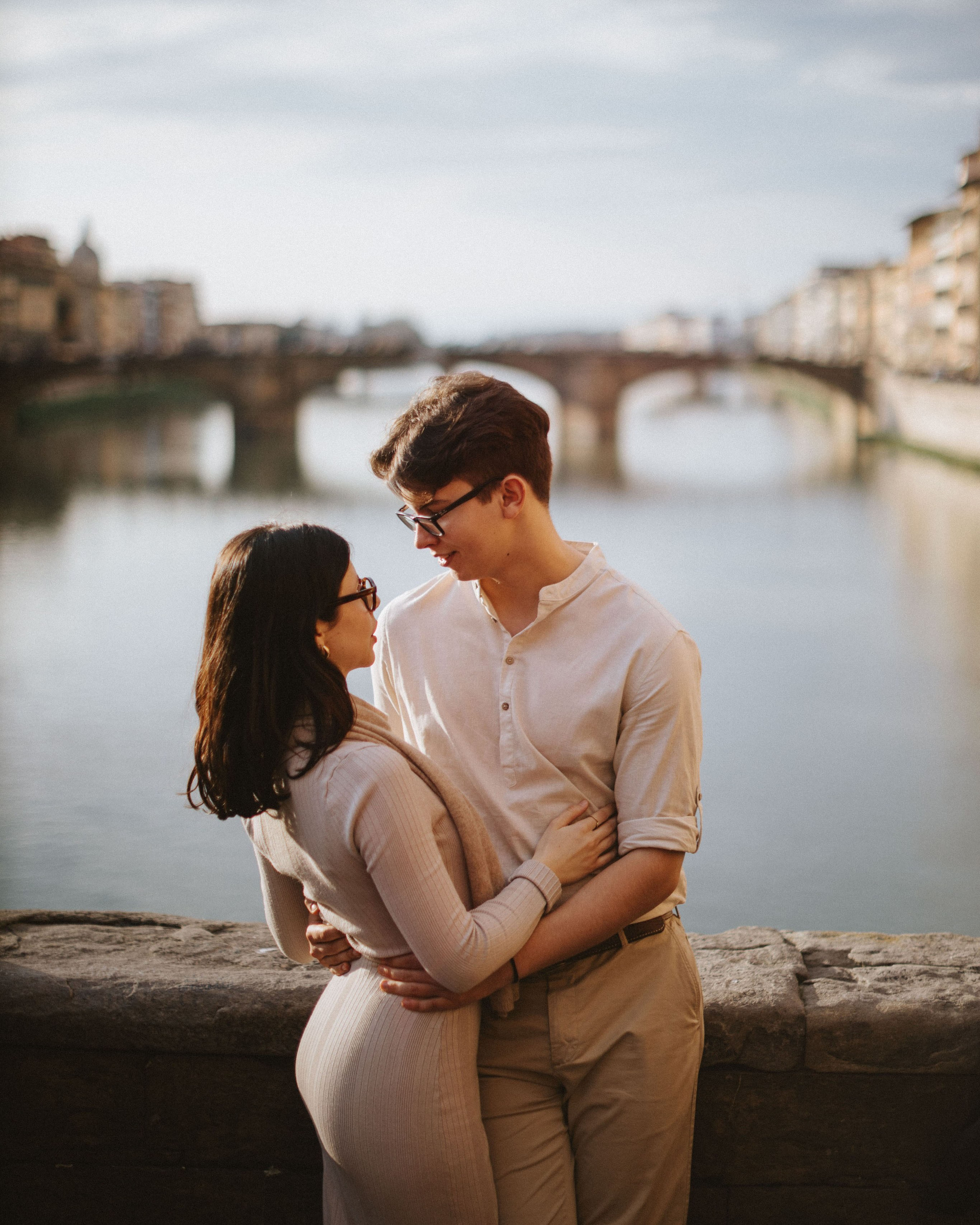 “A couple standing by the river, looking at each other, with the historic Ponte Vecchio bridge in the background.”