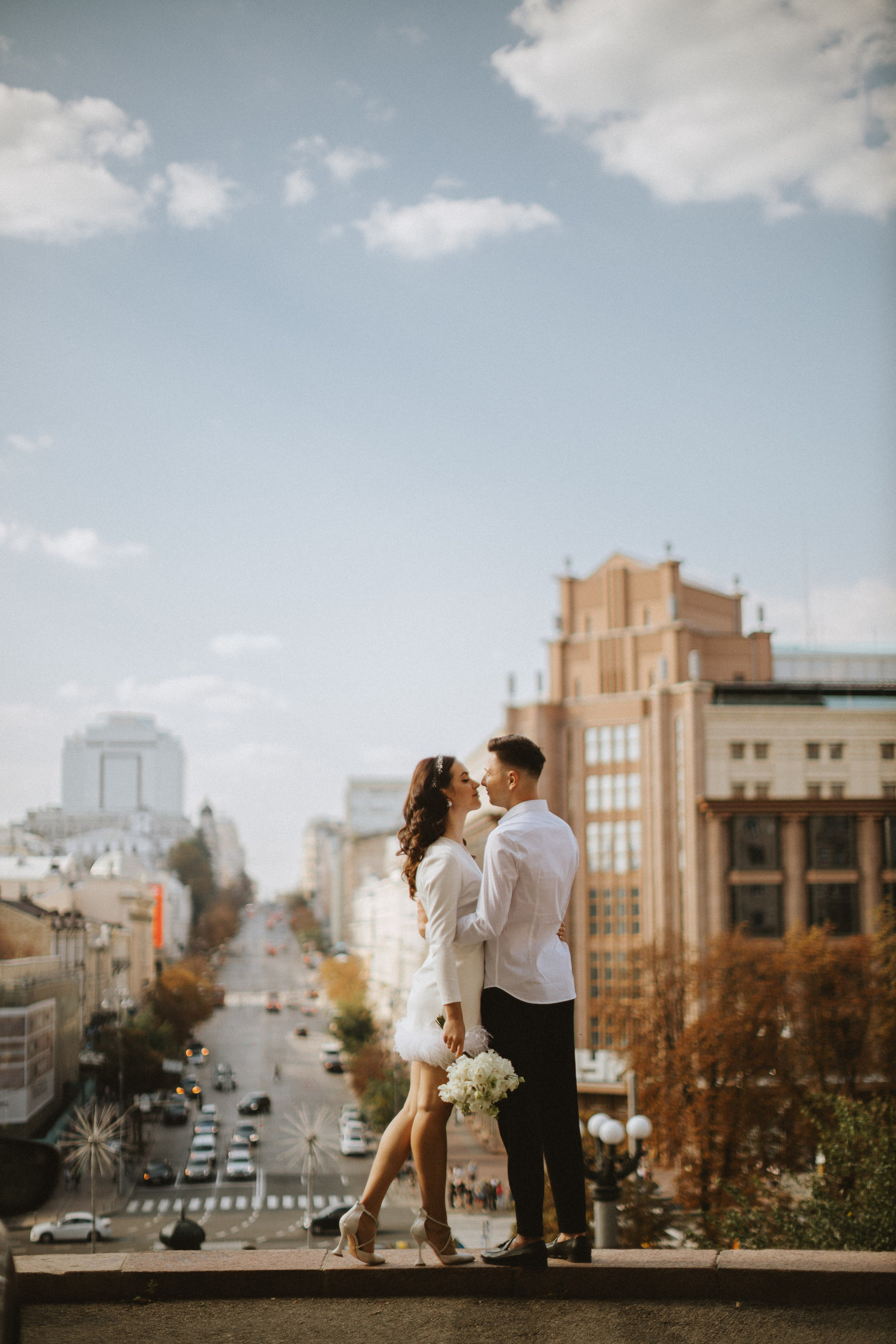 “Una coppia condivide un dolce bacio mentre si trova su un cornicione che domina un bellissimo paesaggio urbano. La sposa, vestita con un elegante completo bianco con accenti di piume, tiene in mano un bouquet di fiori bianchi. Lo sposo, in camicia bianca e pantaloni neri, la abbraccia con un sorriso gioioso. Lo sfondo delle strade e degli edifici della città aggiunge un'atmosfera vibrante e urbana a questo momento intimo”.